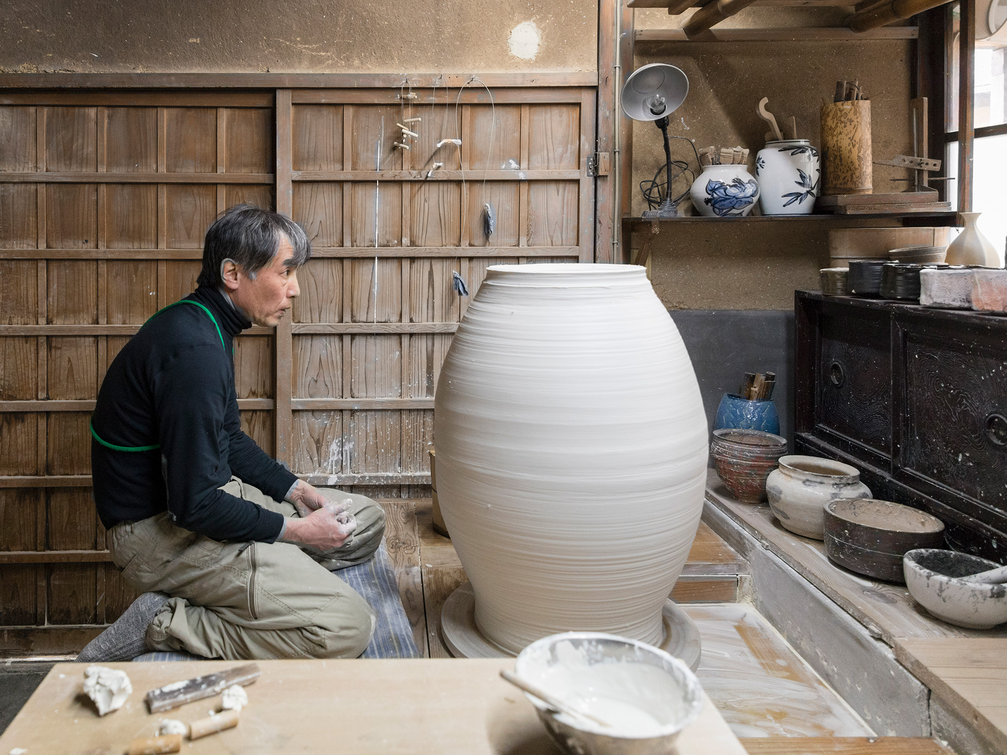 Artist Takahiro Kondo kneeling at the wheel while forming an oversized clay pot inside a traditional...
