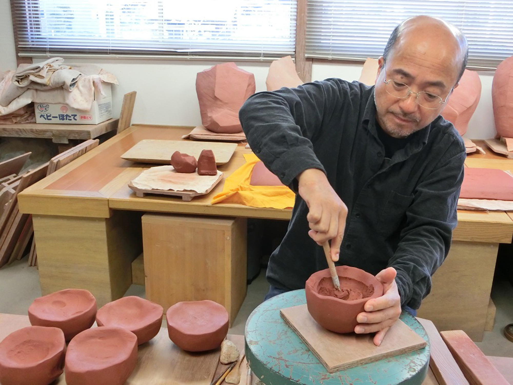 Ceramic artist Masanao Kaneta carving the interior of a hand-built red-clay bowl in a studio filled...