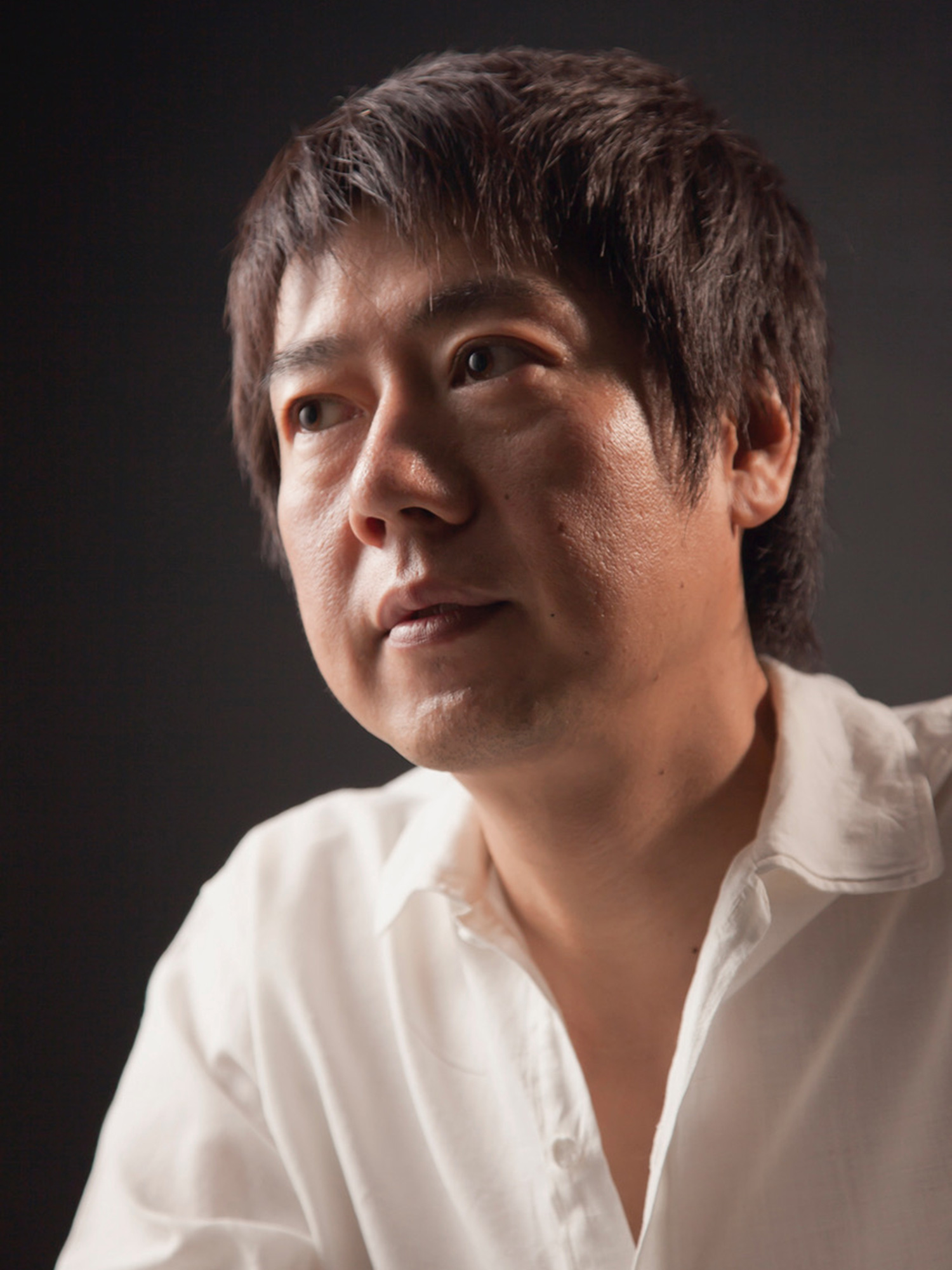 Portrait of Japanese artist Shunichi Yabe in a white shirt photographed against a dark studio background...