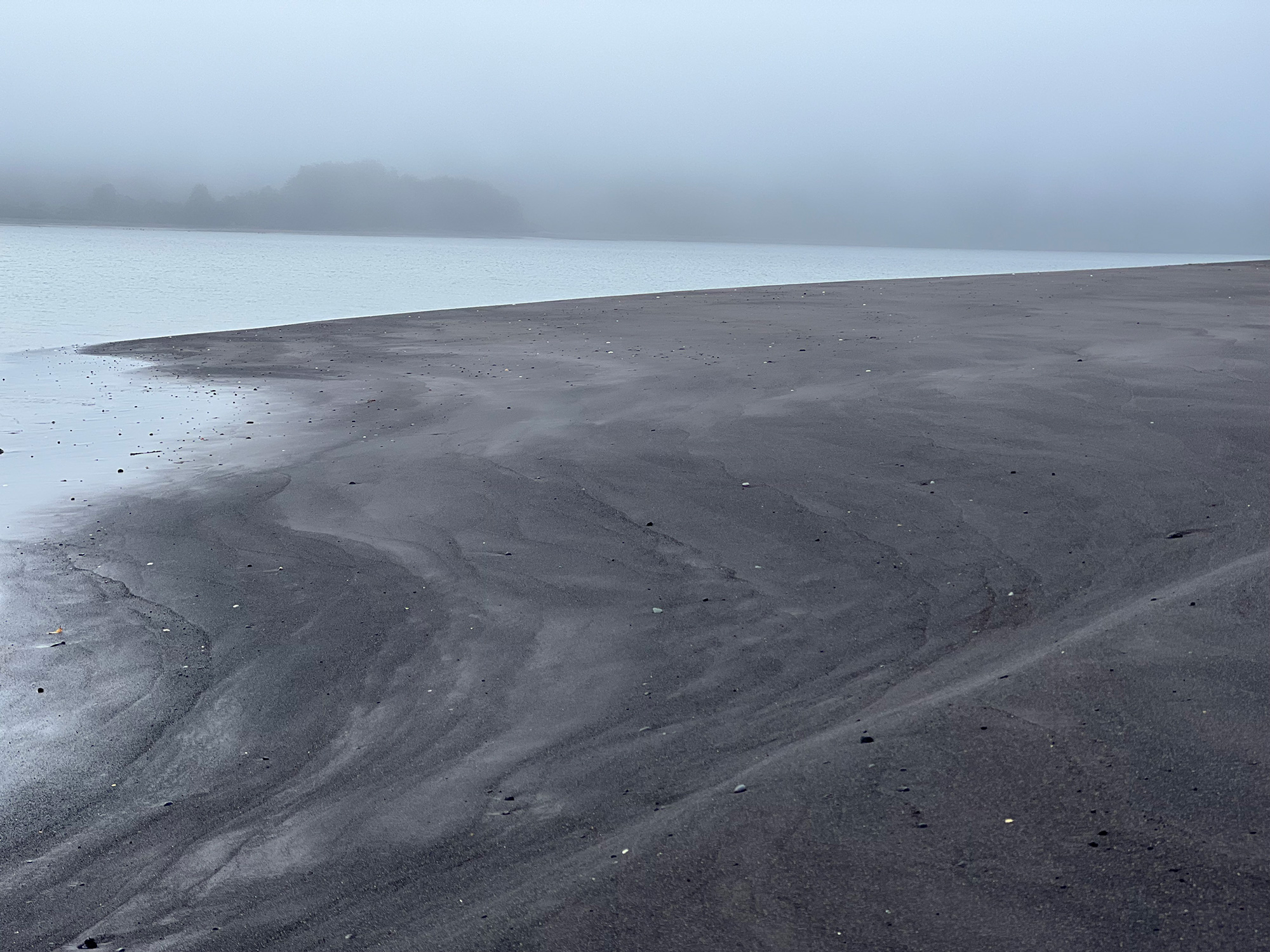 Misty coastal scene at Tic Toc showing smooth black volcanic sand meeting the calm, fog-covered...