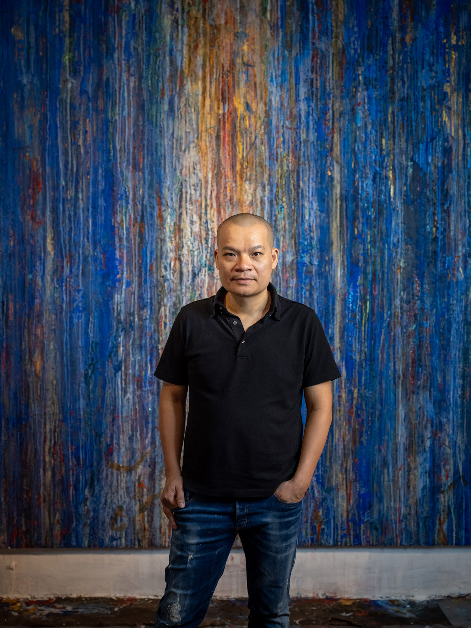 Contemporary artist Ha Manh Thang in a studio posed before a tall, textured abstract painting in...