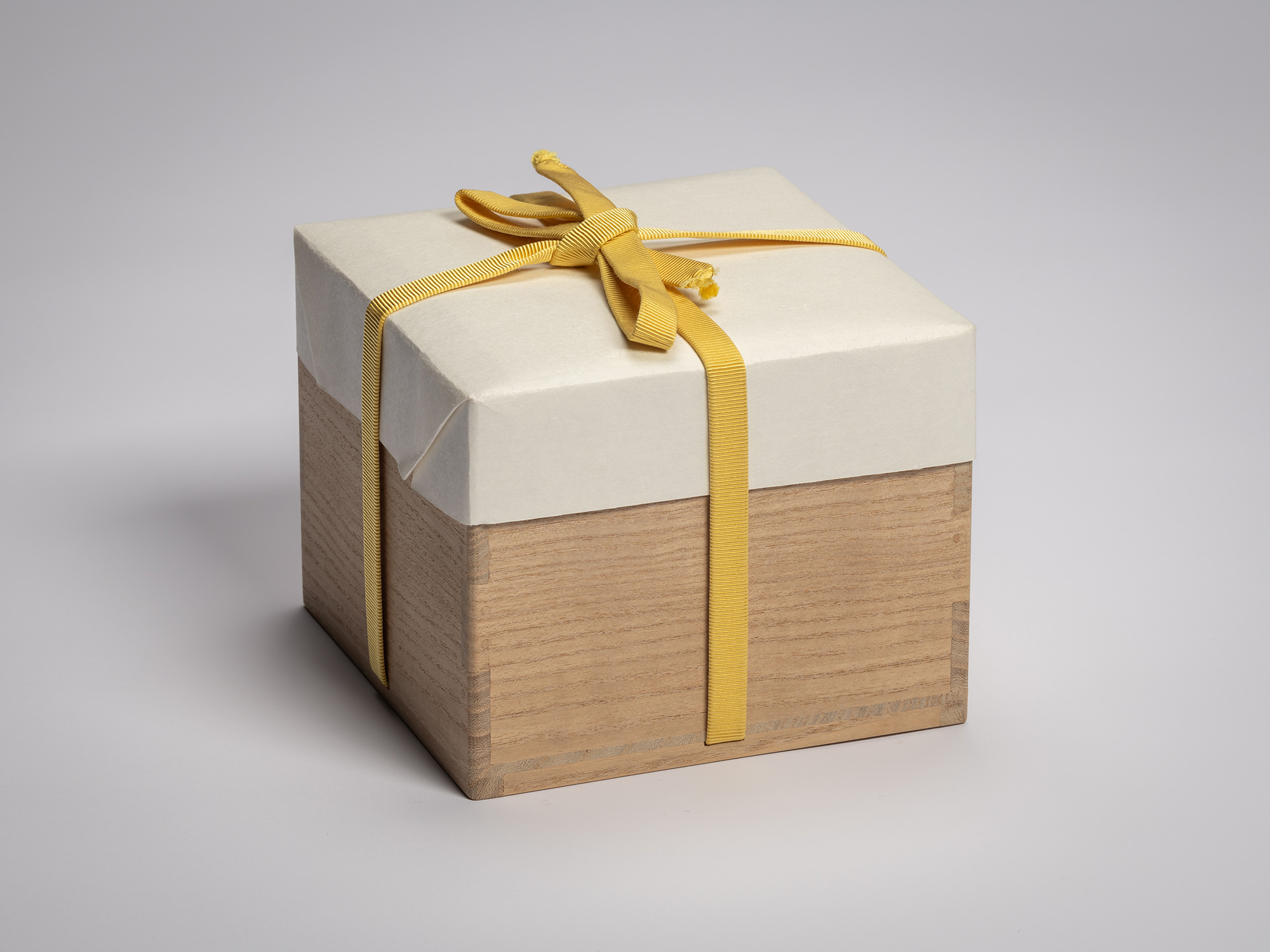 Square wooden tomobako (storage box), shown with its mustard cloth carrying strap. The box is half-wrapped...