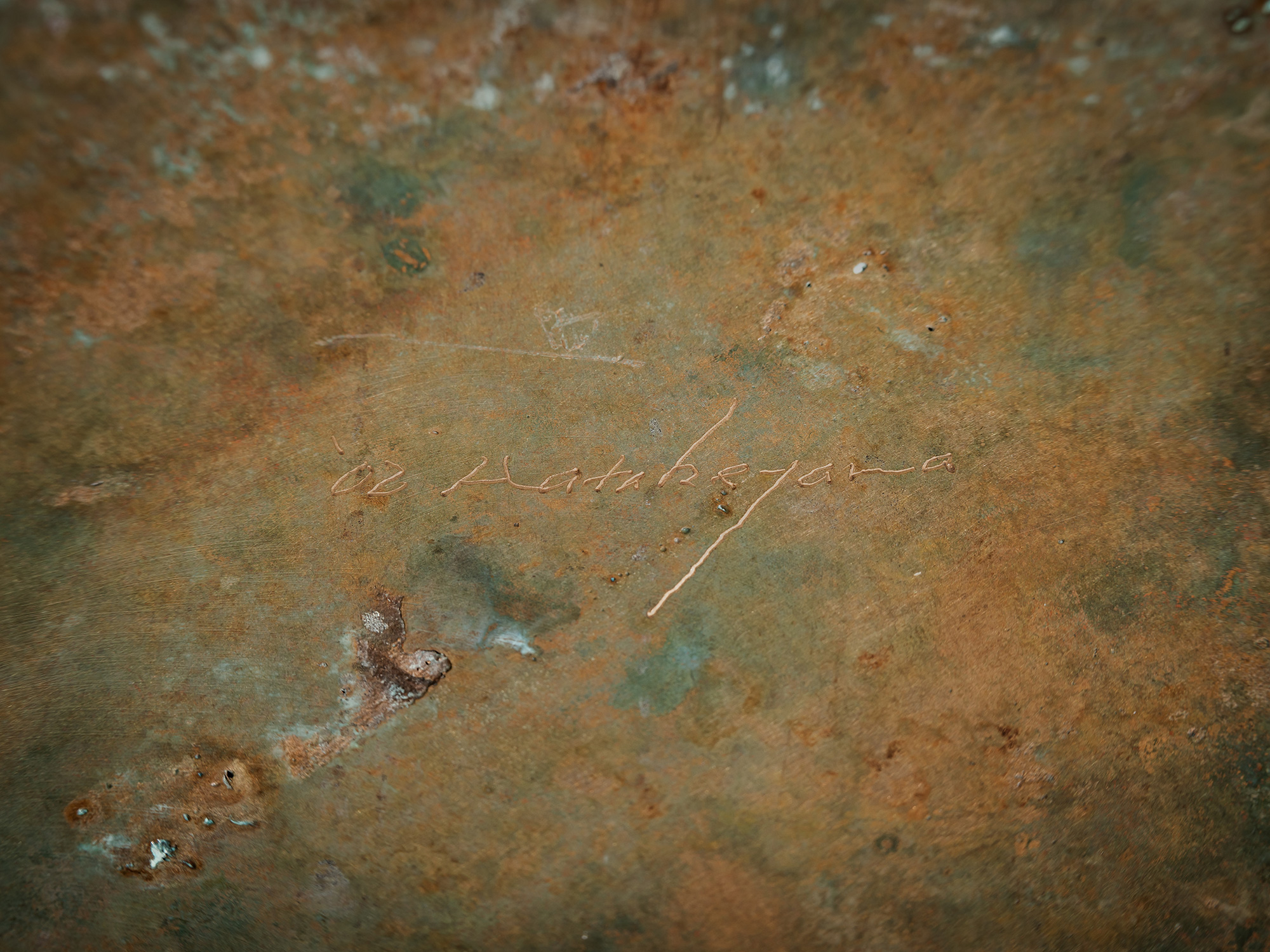 Close-up photograph of the incised signature of Koji Hatakeyama, scratched into a bronze surface....
