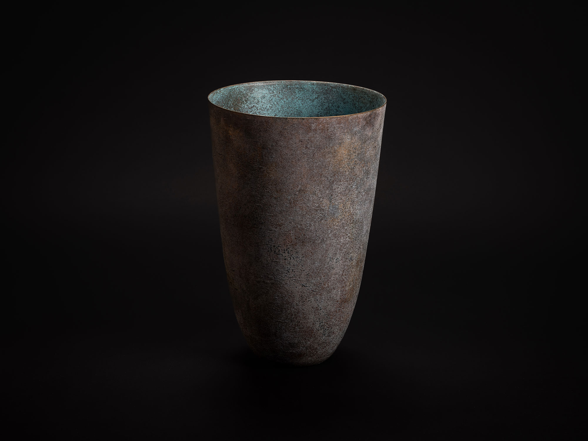 Tall bronze vase by Koji Hatakeyama, displayed against a black background,with a tapered cylindrical...