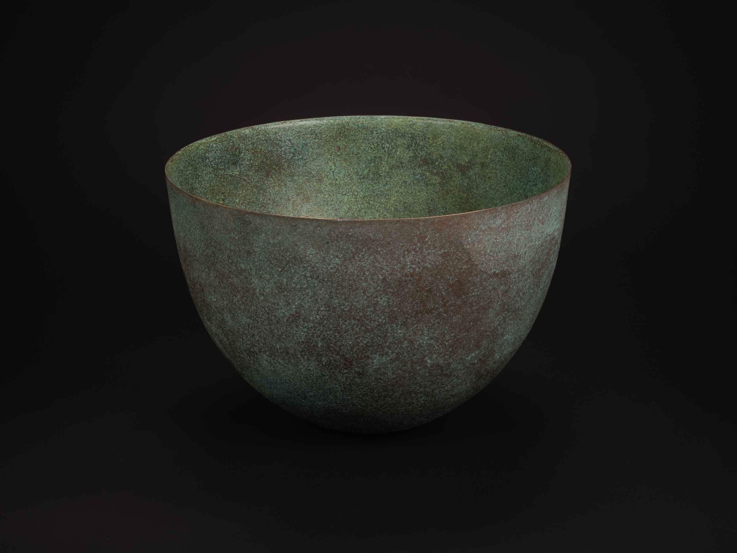 Rounded bronze vessel titled Bronze No. 17 by Hatakeyama set against a dark background, with a...