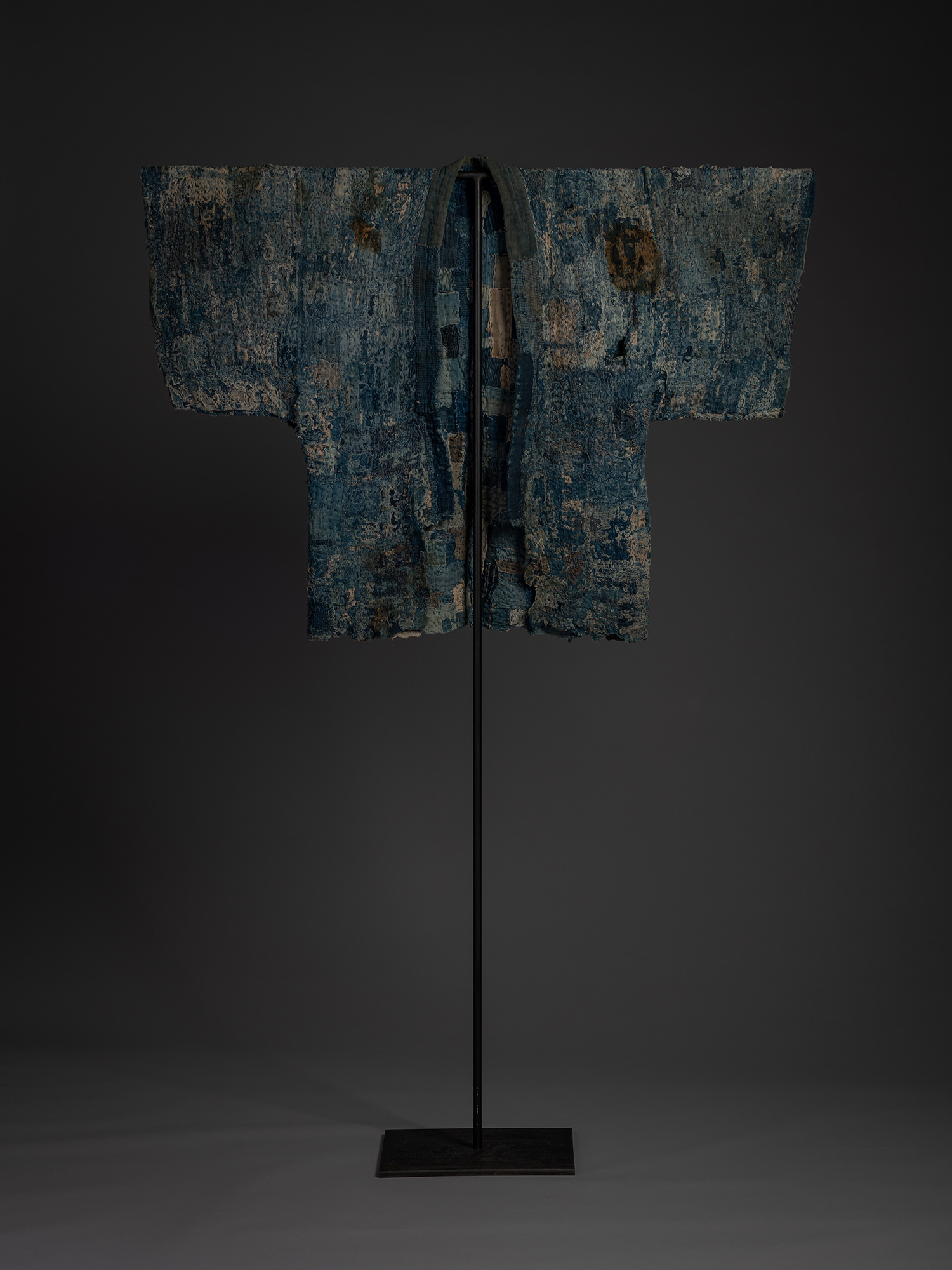 Full installation view of an indigo-dyed boro hanten mounted on a tall black metal stand with square...