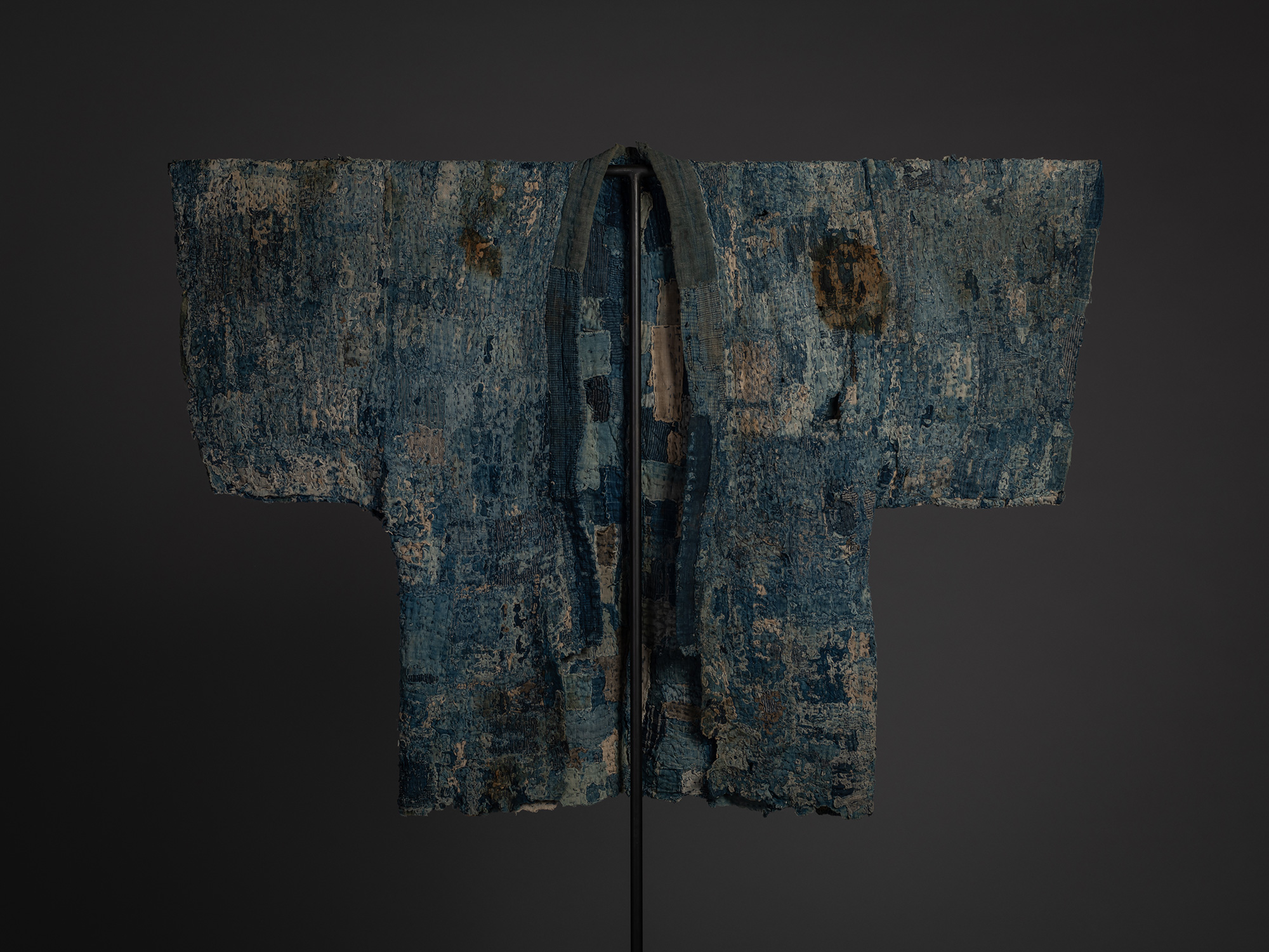 Frontal view of a Japanese boro hanten mounted on a black stand against a dark background. The...