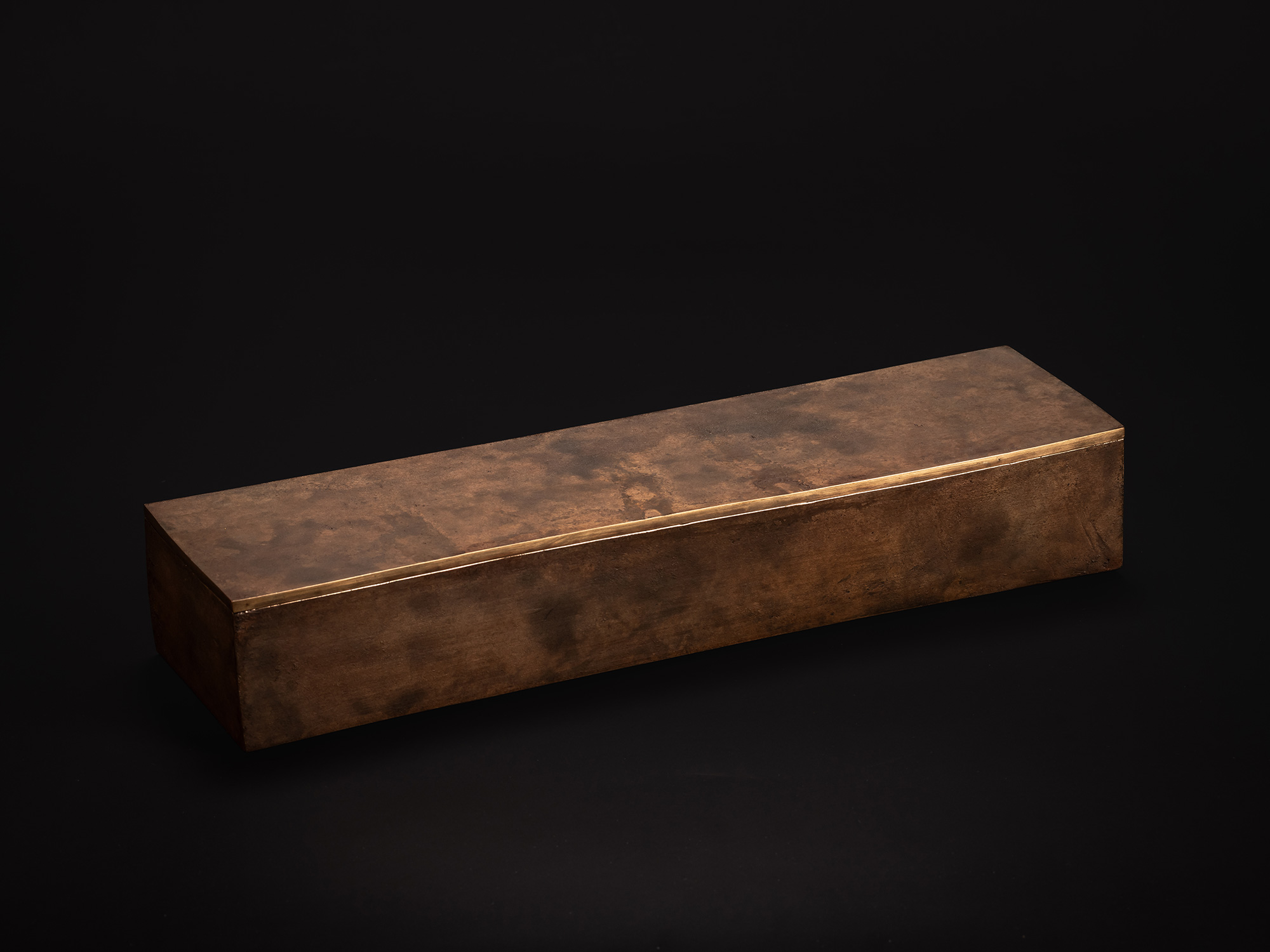 Three-quarter view of a long rectangular bronze box with a slightly arched lid. The brown patinated...