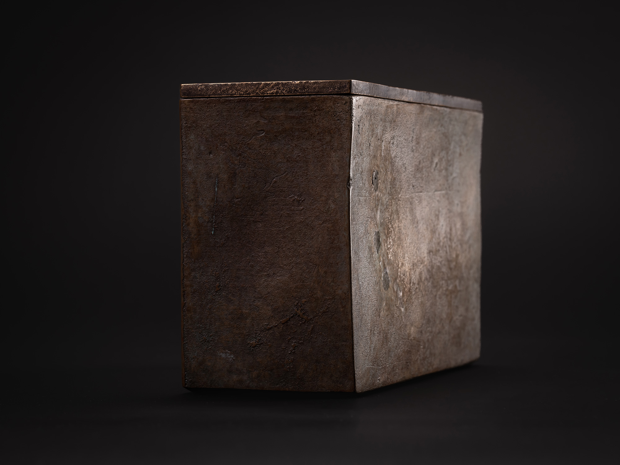Three-quarter view of a rectangular bronze box with a fitted flat lid. The exterior surface shows...