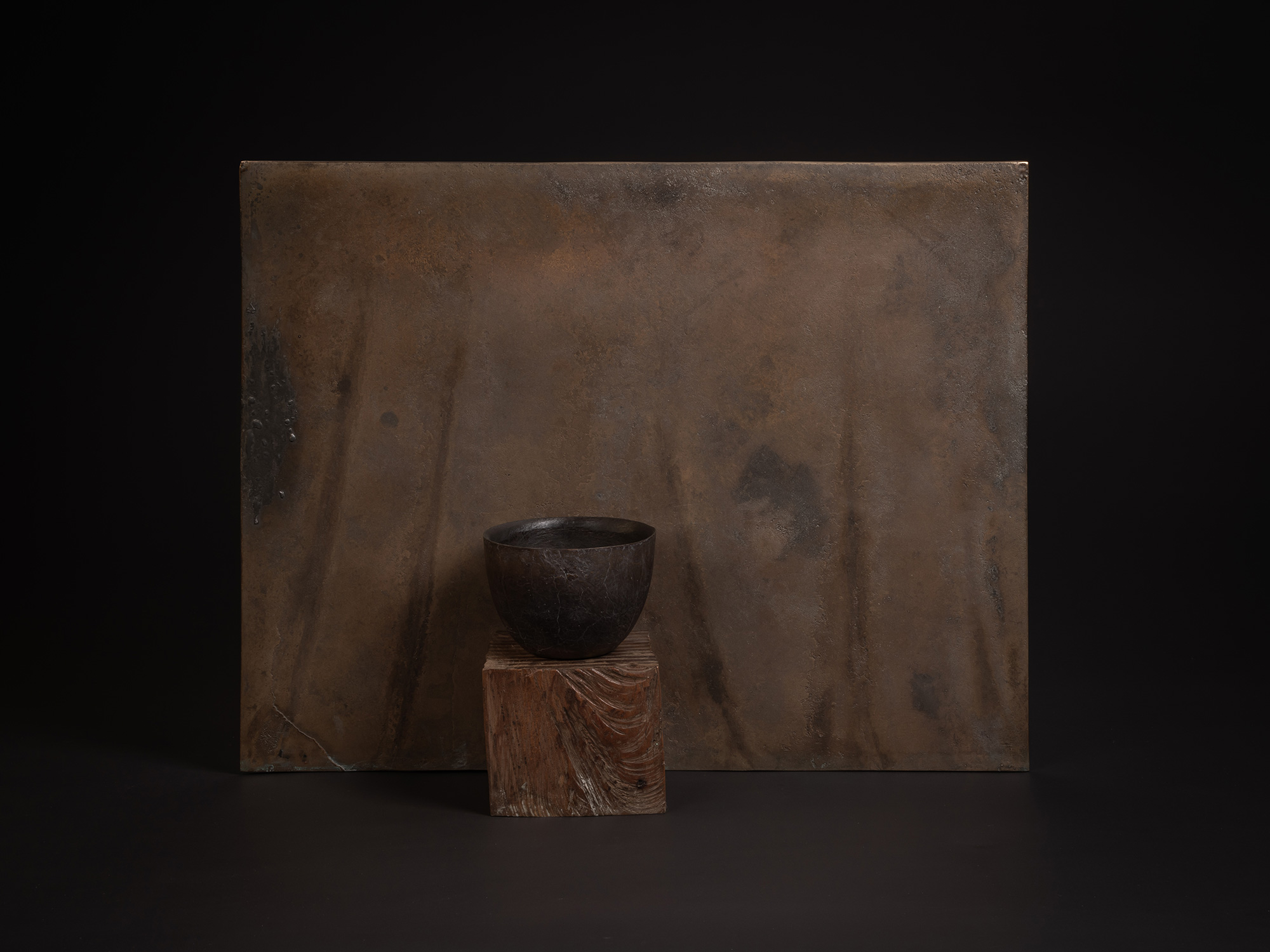 A small dark bronze vessel by Koji Hatakeyama sits on a carved wooden block before a large rectangular...