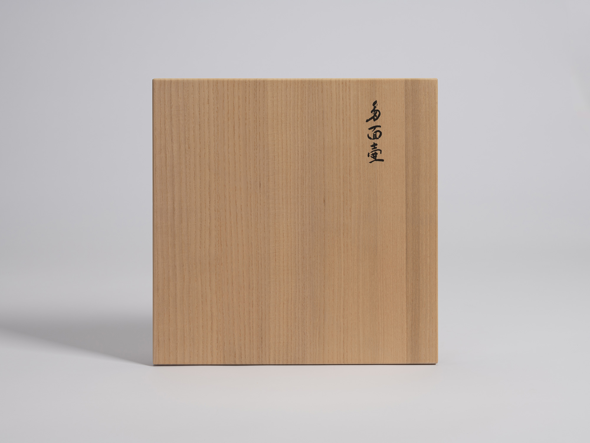 A square, fine-grained paulownia wood box lid (tomobako) viewed straight-on against a seamless...