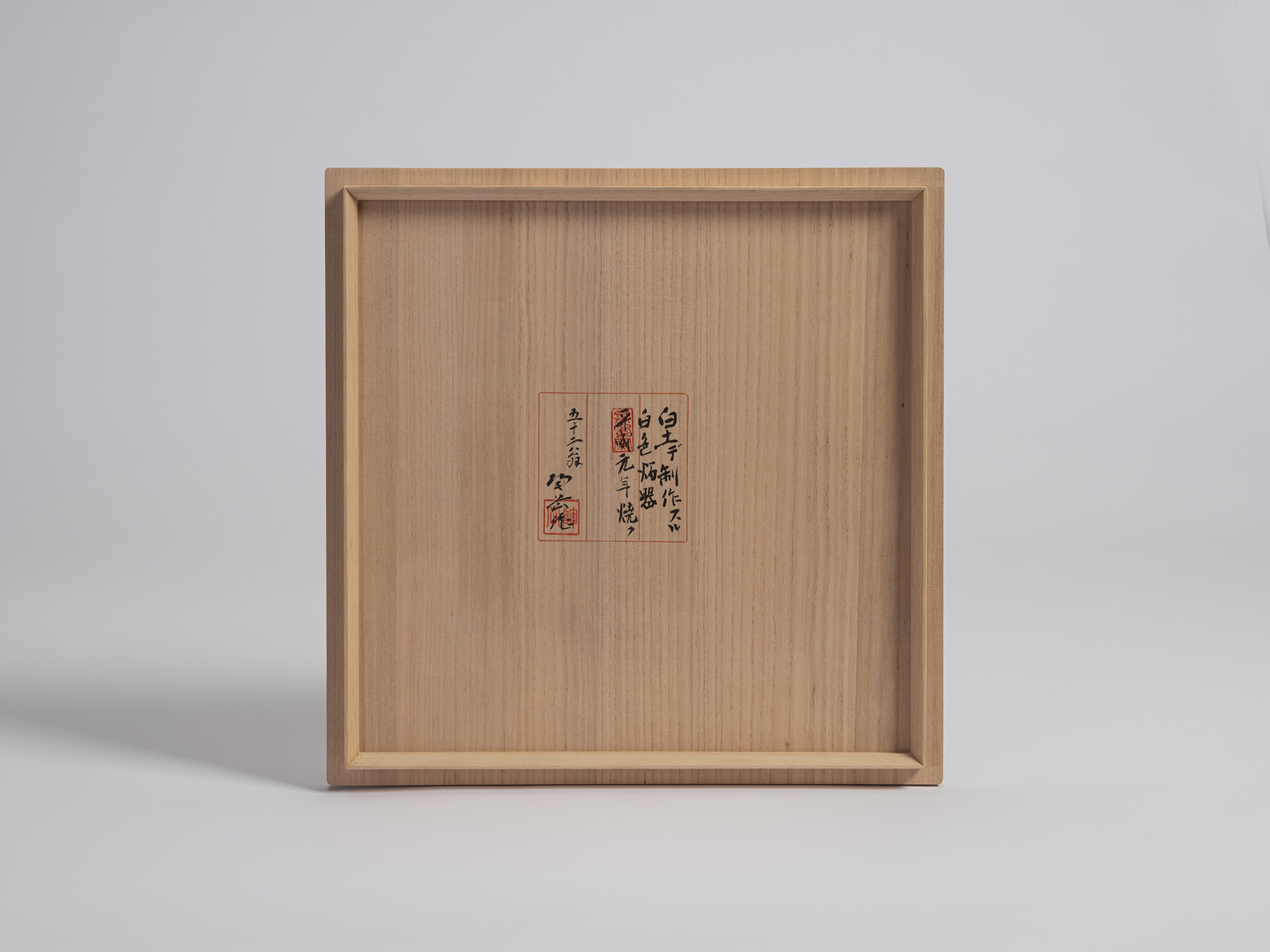 The square underside of a paulownia wood box lid (tomobako), viewed straight-on against a seamless...
