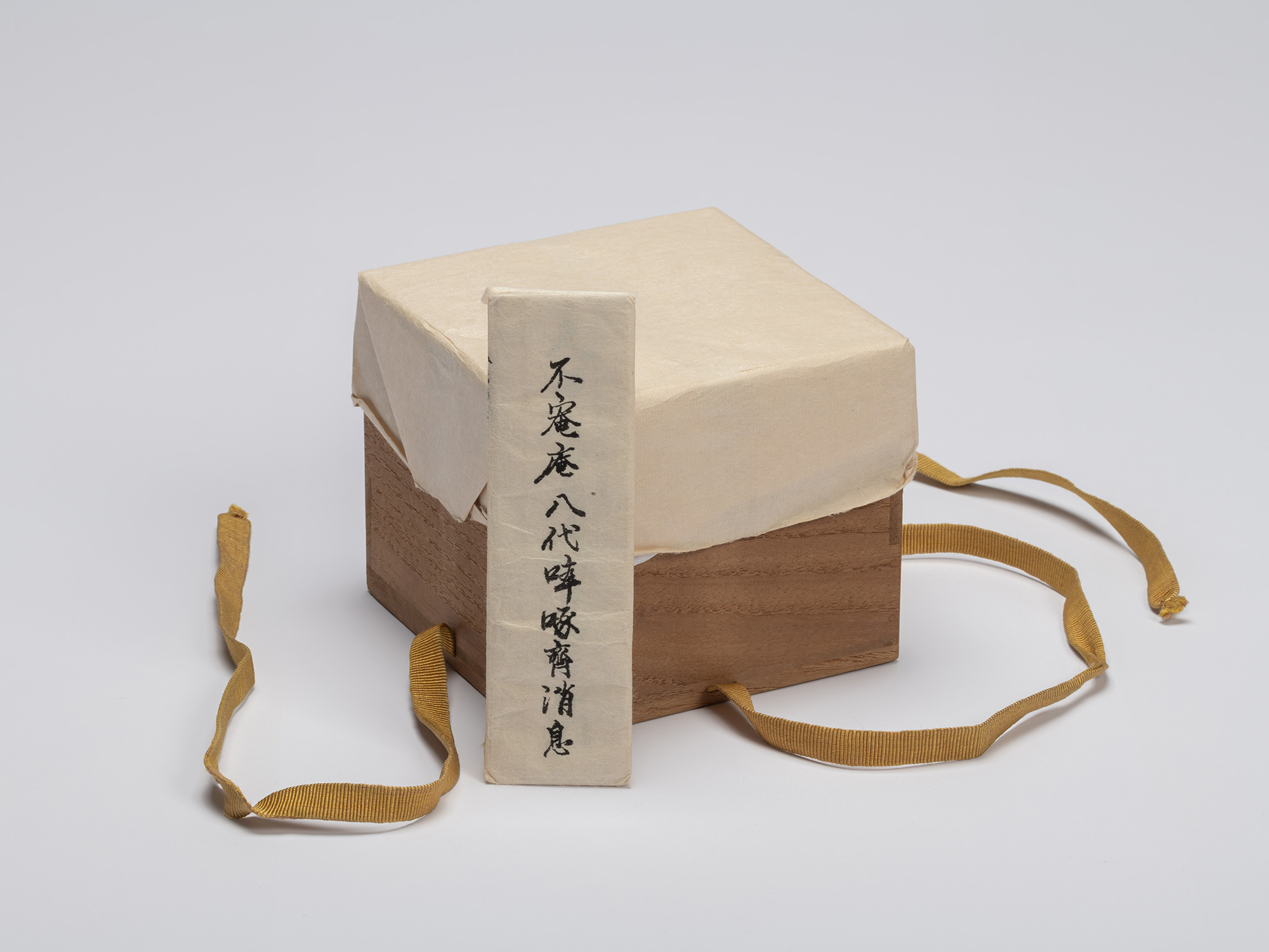 A square, dark paulownia wood storage box (tomobako) sits centered against a seamless light gray...