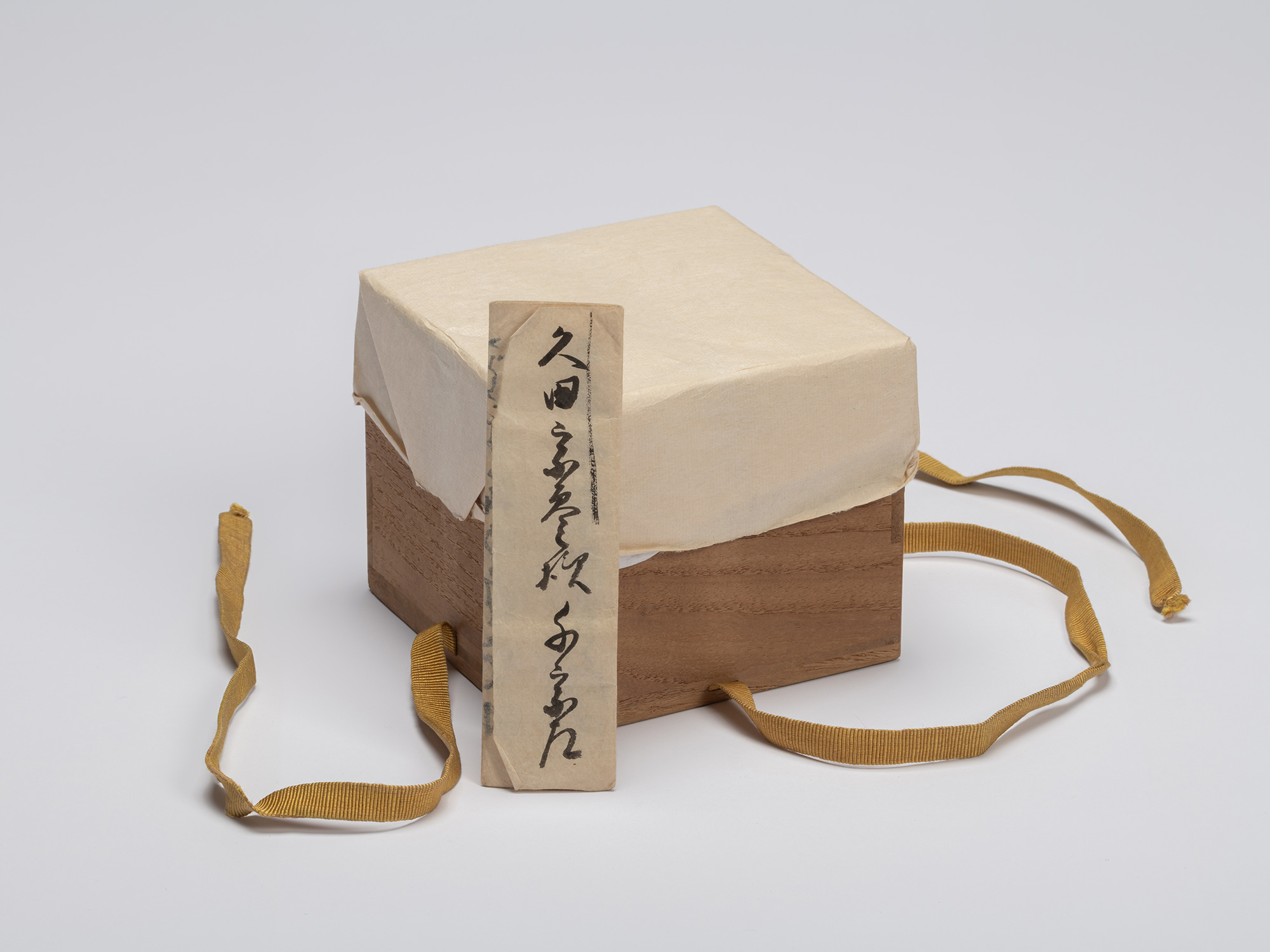 A square, dark paulownia wood storage box (tomobako) is centered against a seamless light gray...