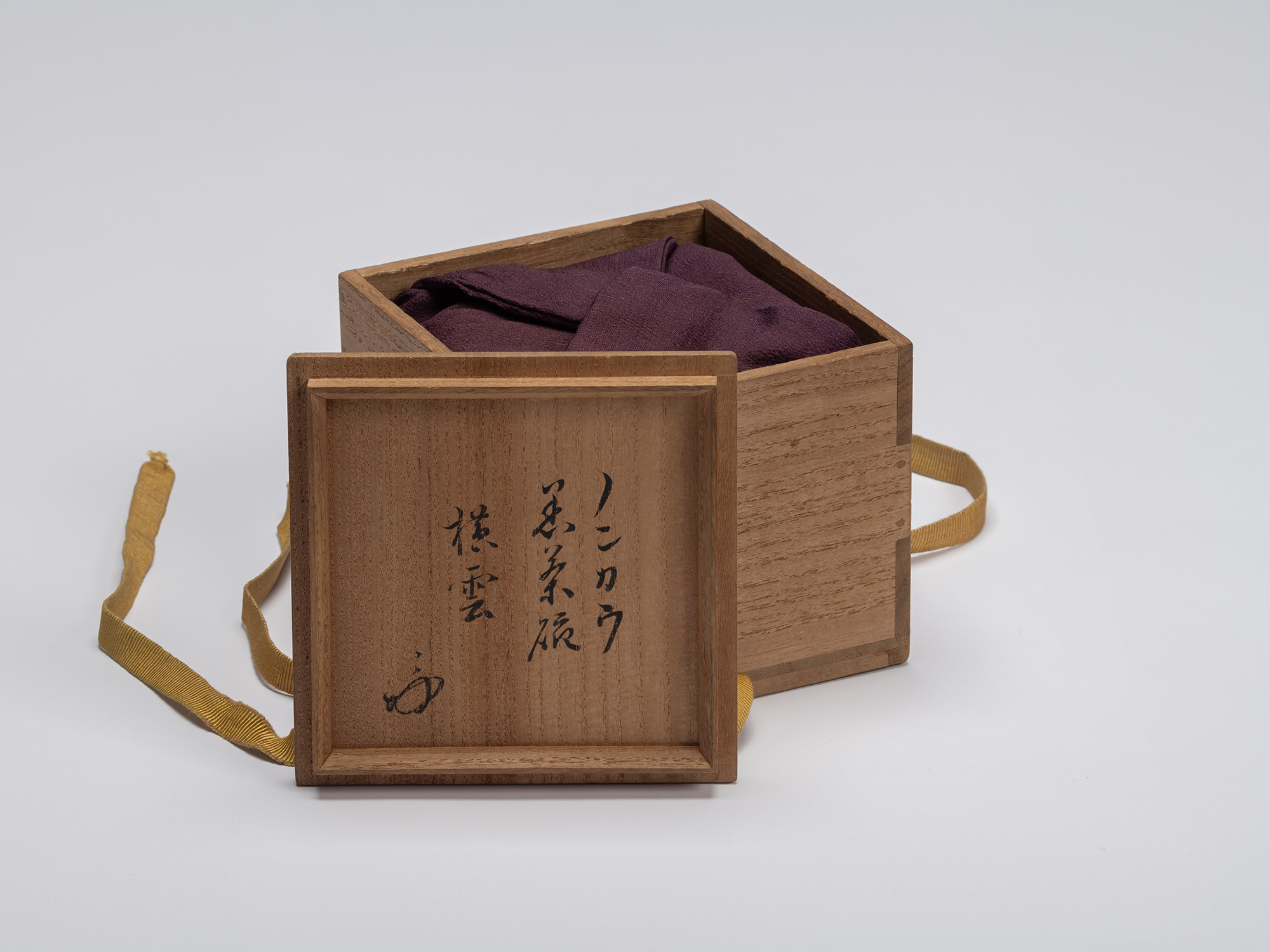 The square, light-colored paulownia wood outer storage box (tomobako) sits open against a seamless...