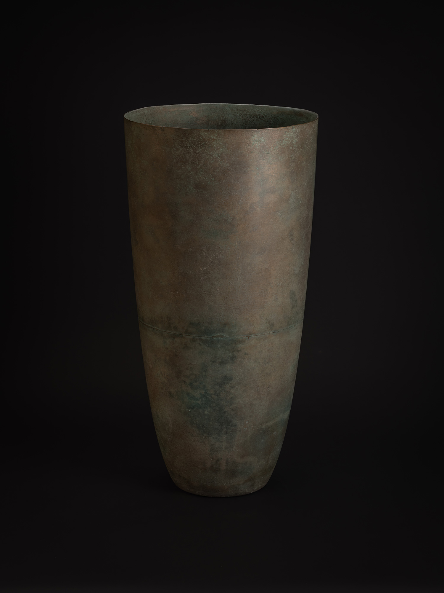 A tall bronze vessel with a broad oval opening and softly rounded contours. The surface features...