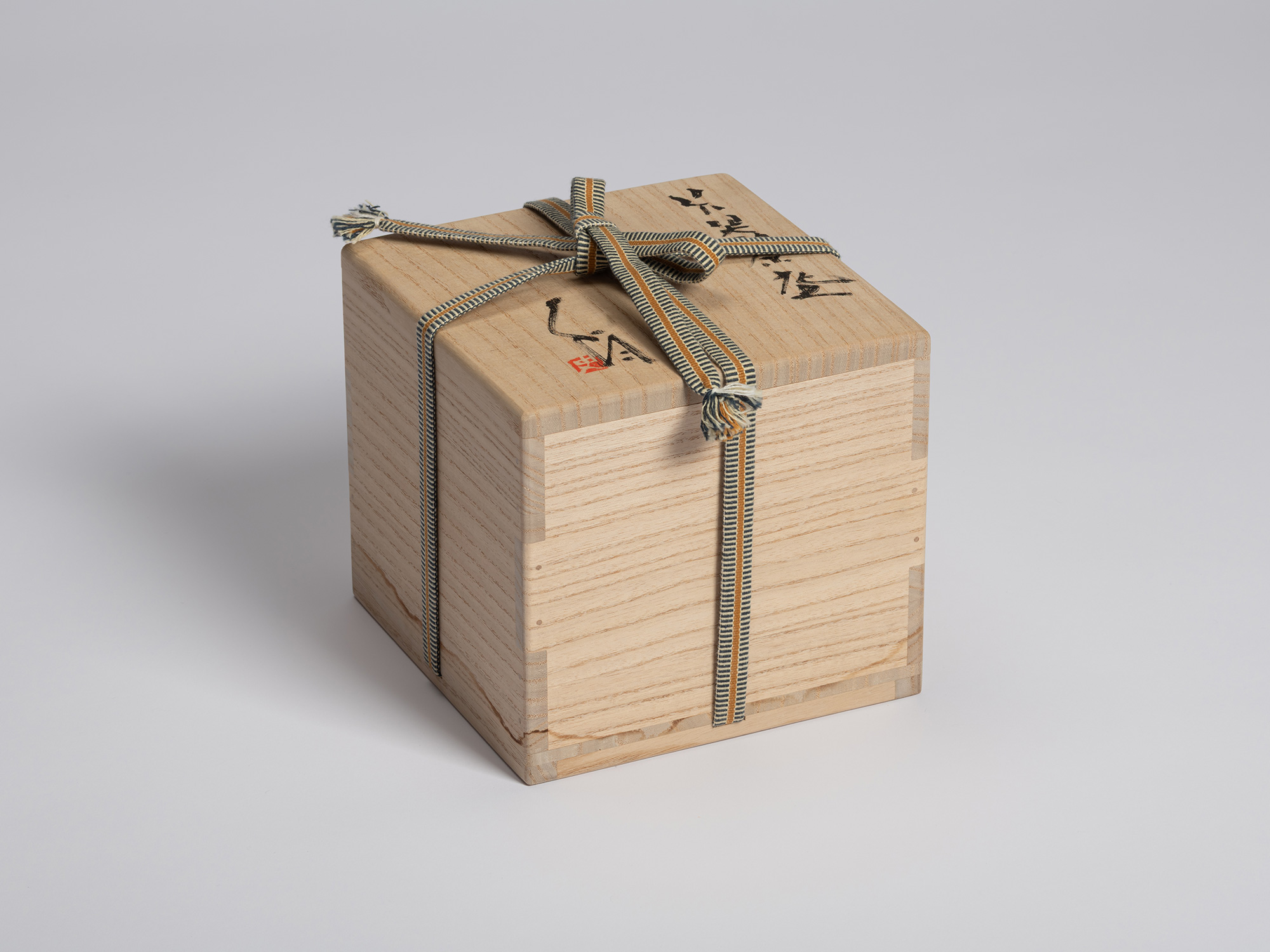 Paulownia wood tomobako secured with a striped braided cord tied across the lid. Black ink calligraphy...