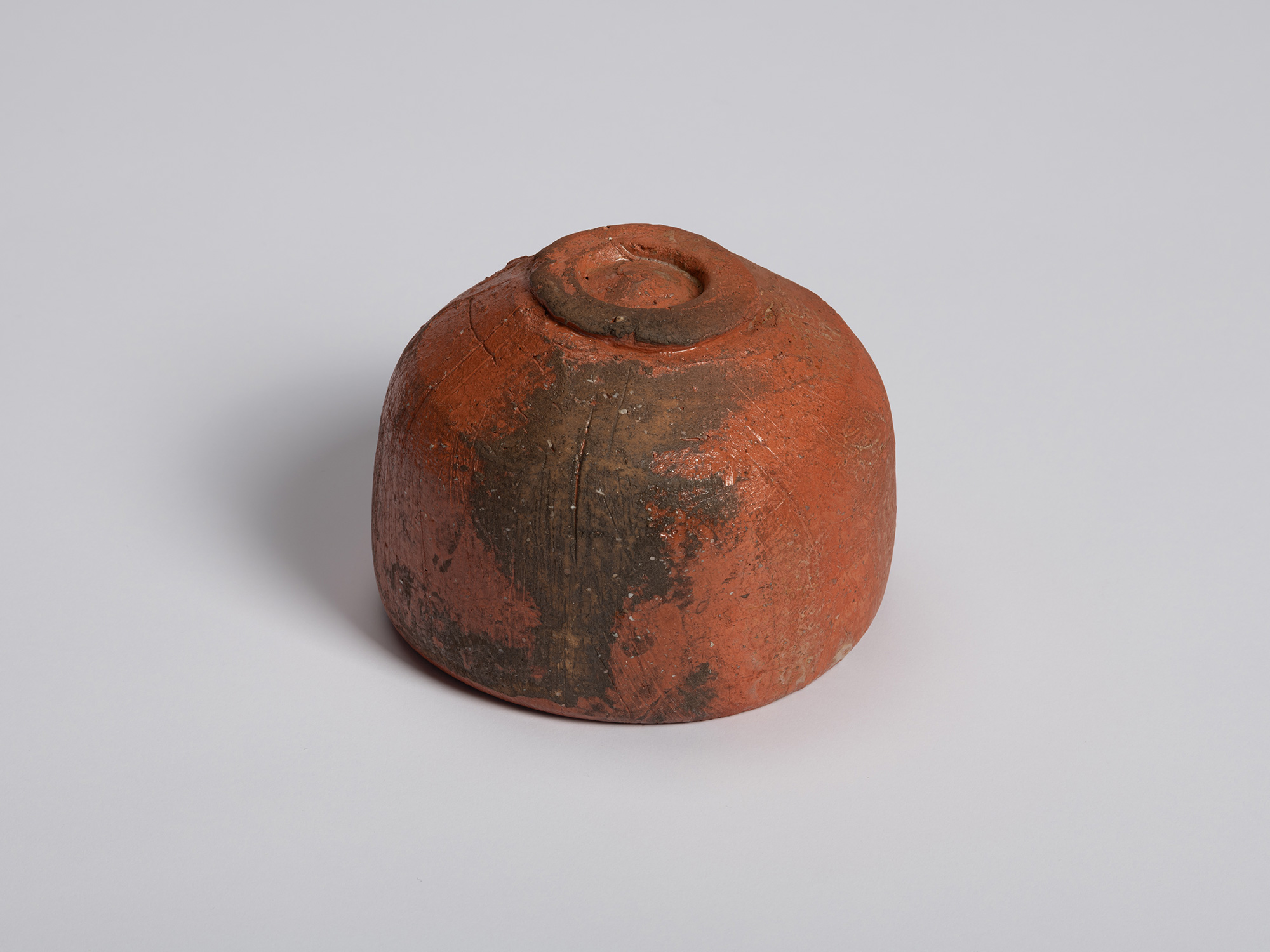 Angled view of the underside of a red Raku tea bowl by Shiro Tsujimura. The raised foot ring projects...