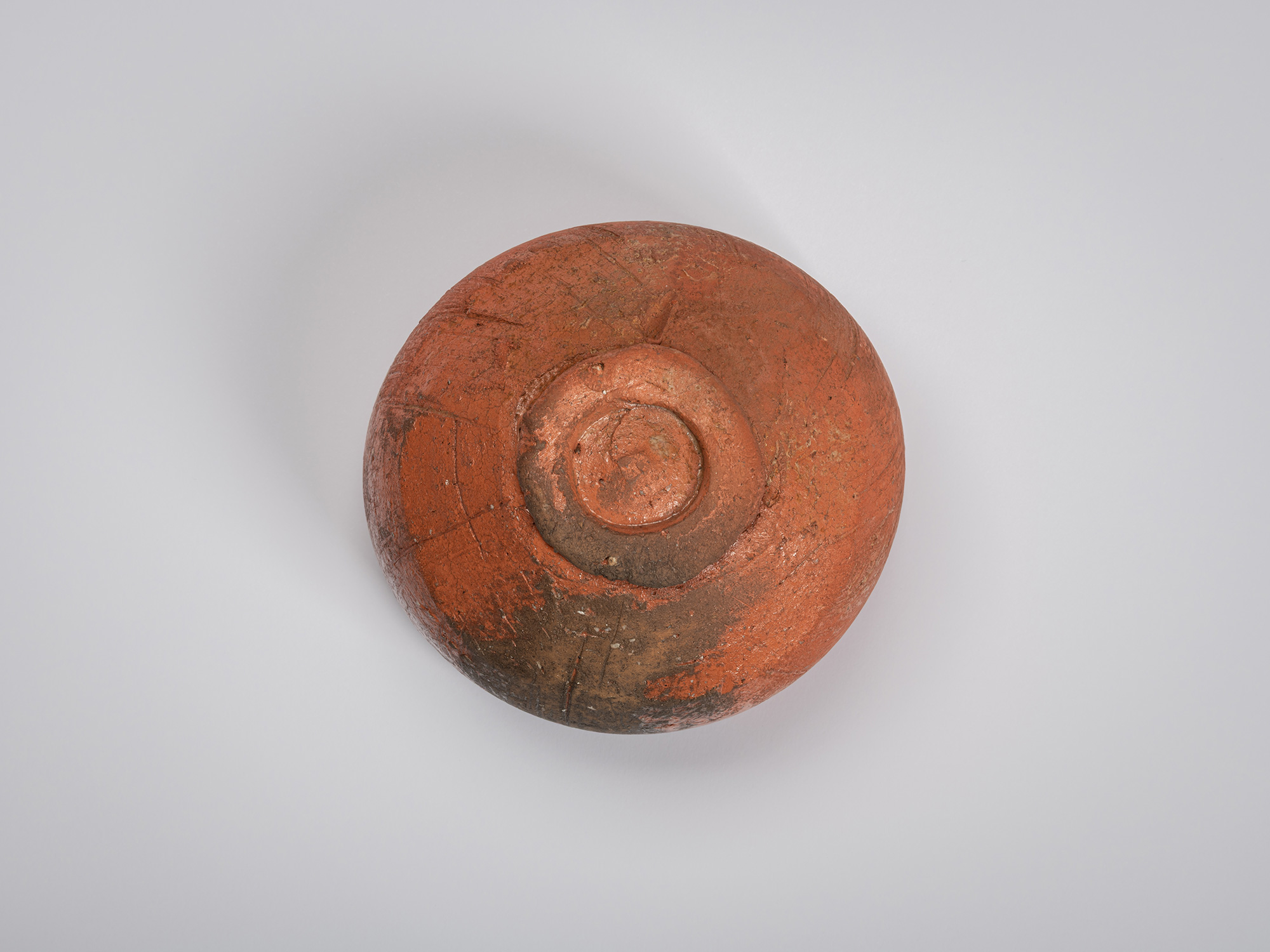 Bottom view of a red Raku tea bowl by Shiro Tsujimura. The underside reveals a small circular foot...