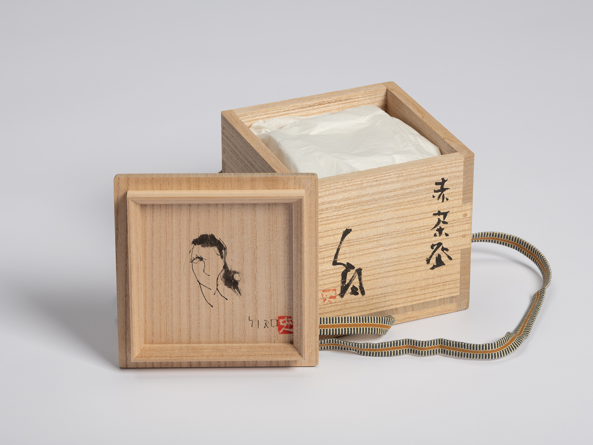 Open wooden tomobako accompanying a red Raku tea bowl by Shiro Tsujimura. The kiri wood box is...