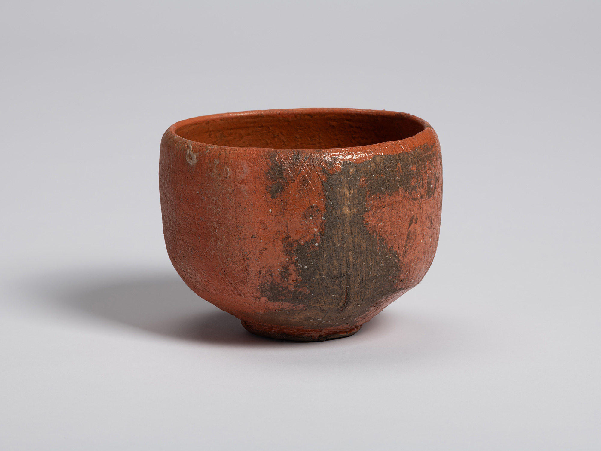 View of a red Raku tea bowl by Shiro Tsujimura. A dark, irregular firing mark extends vertically...