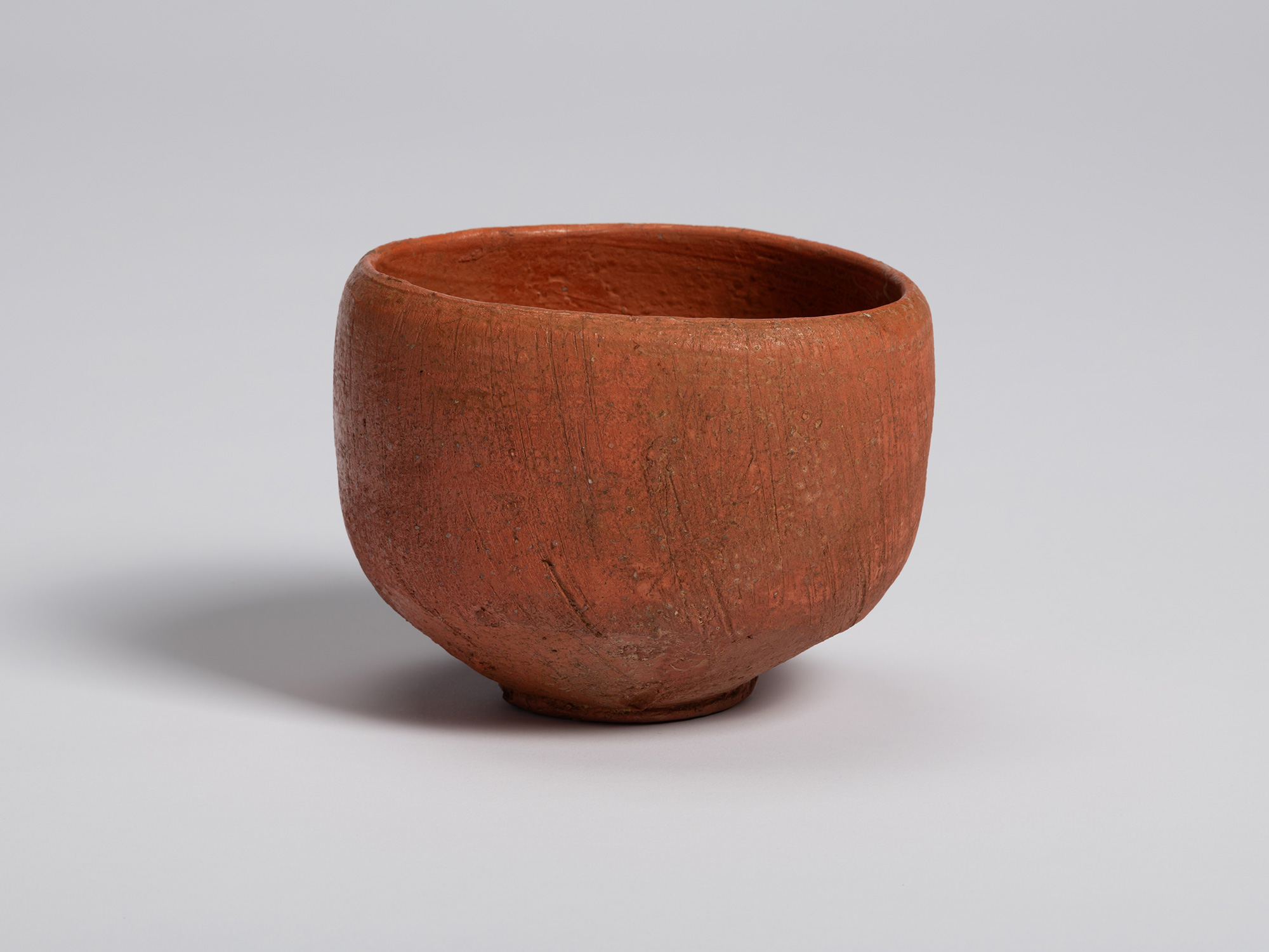 Red Raku tea bowl by Shiro Tsujimura showing a largely uninterrupted red surface. Vertical tool...