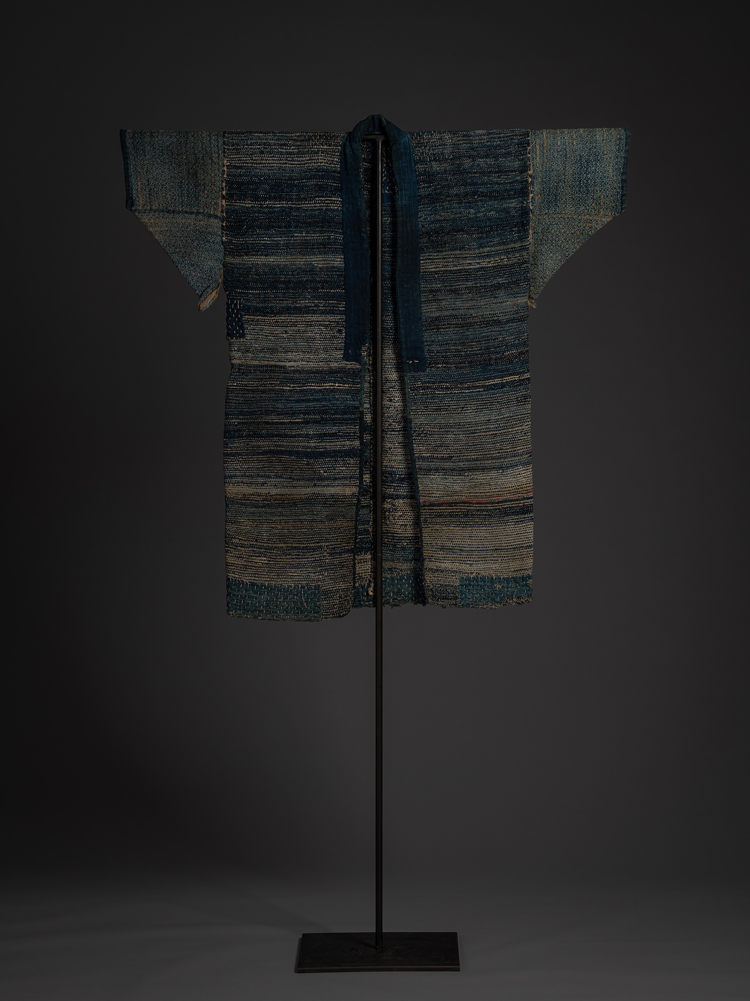 Front view of the Japanese sakiori hanten displayed on a stand. The garment features a long indigo...