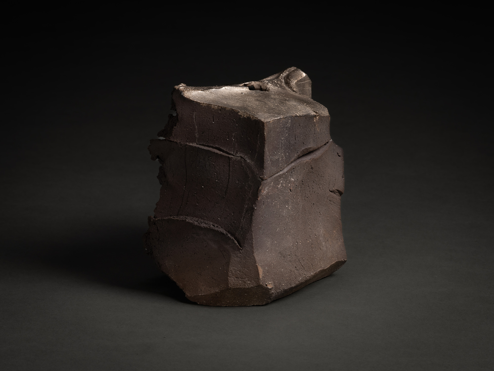 Profile of the sculpture highlighting its angular, block-like form. The smooth, dark clay surfaces...