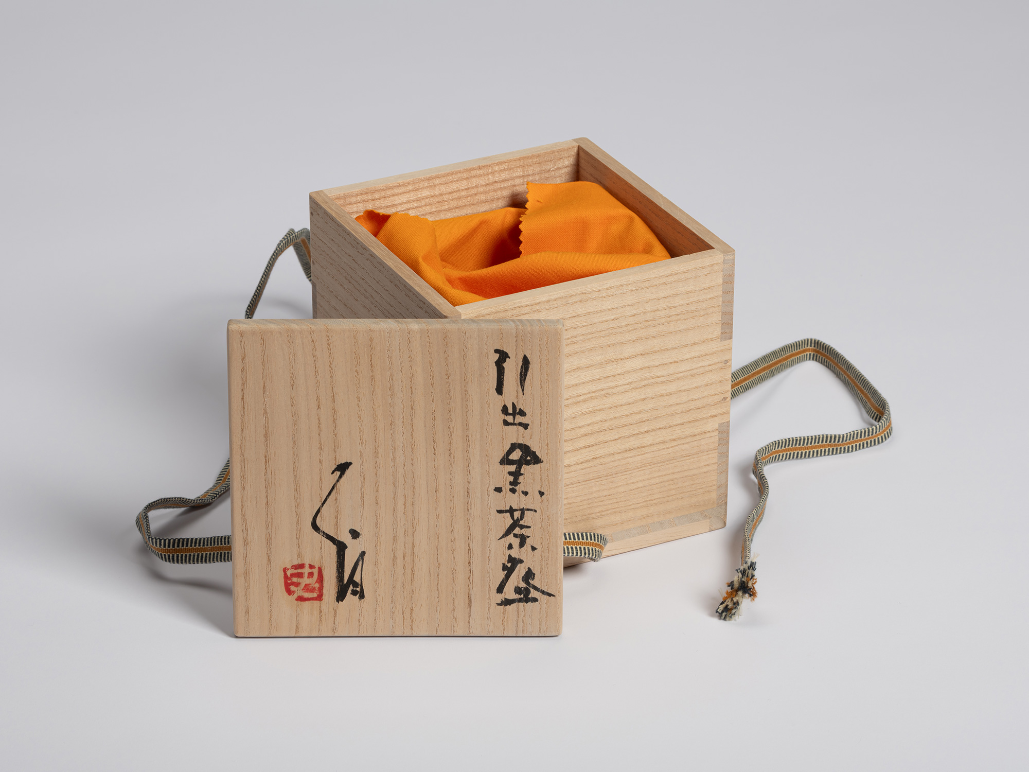 Wooden tomobako displayed with the lid set in front of the open box. Bright orange cloth used to...