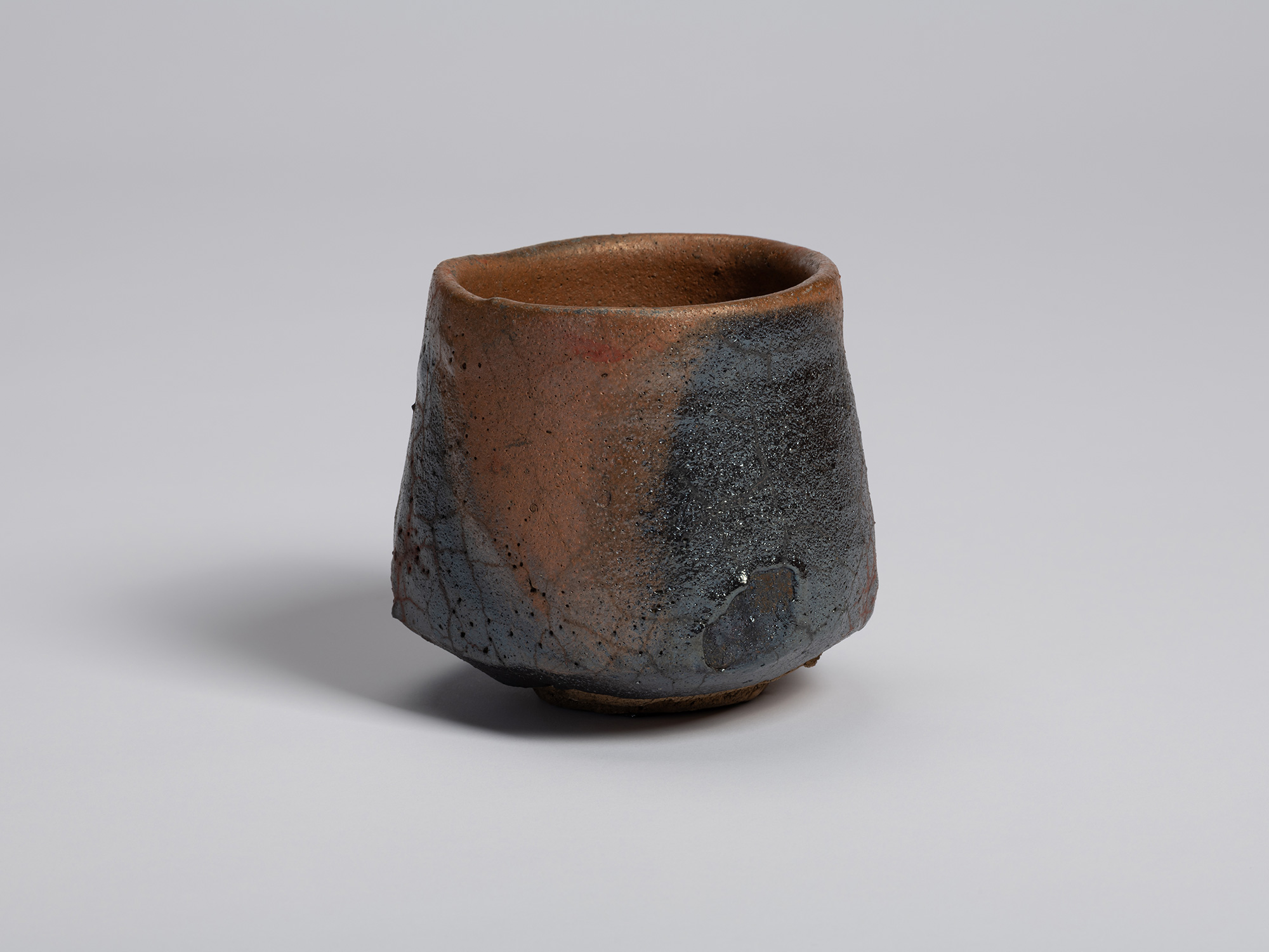 View of the tea bowl showing its gently faceted profile and uneven rim. Areas of black glaze blend...