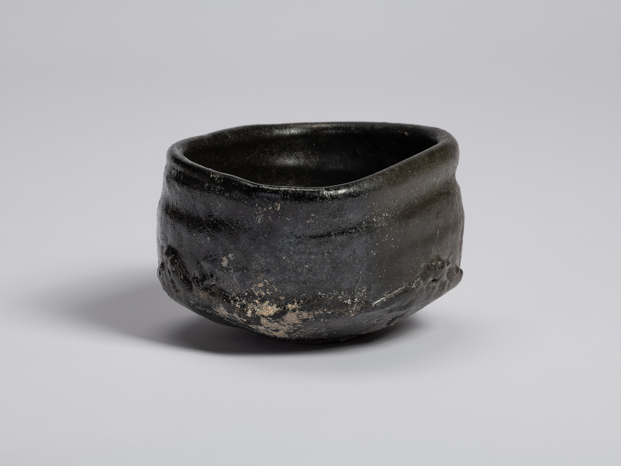View of a black tea bowl by Shiro Tsujimura showing its asymmetrical form and uneven rim. The lustrous...