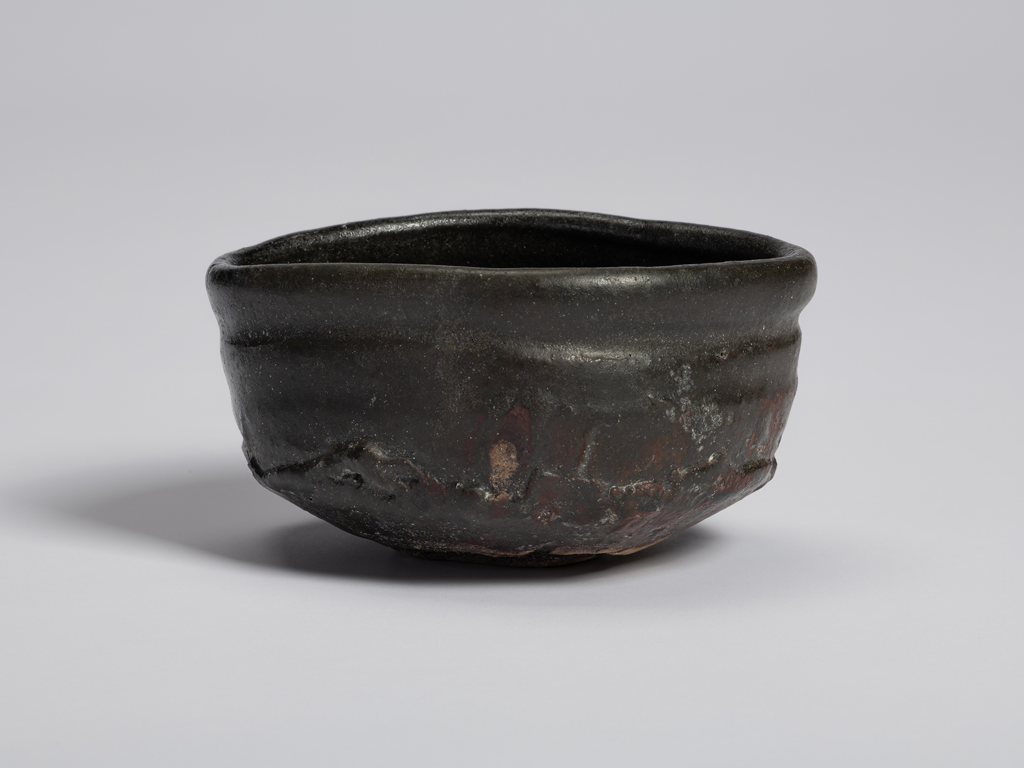 View of a black tea bowl by Shiro Tsujimura. The vessel features a broad, rounded body with a subtly...
