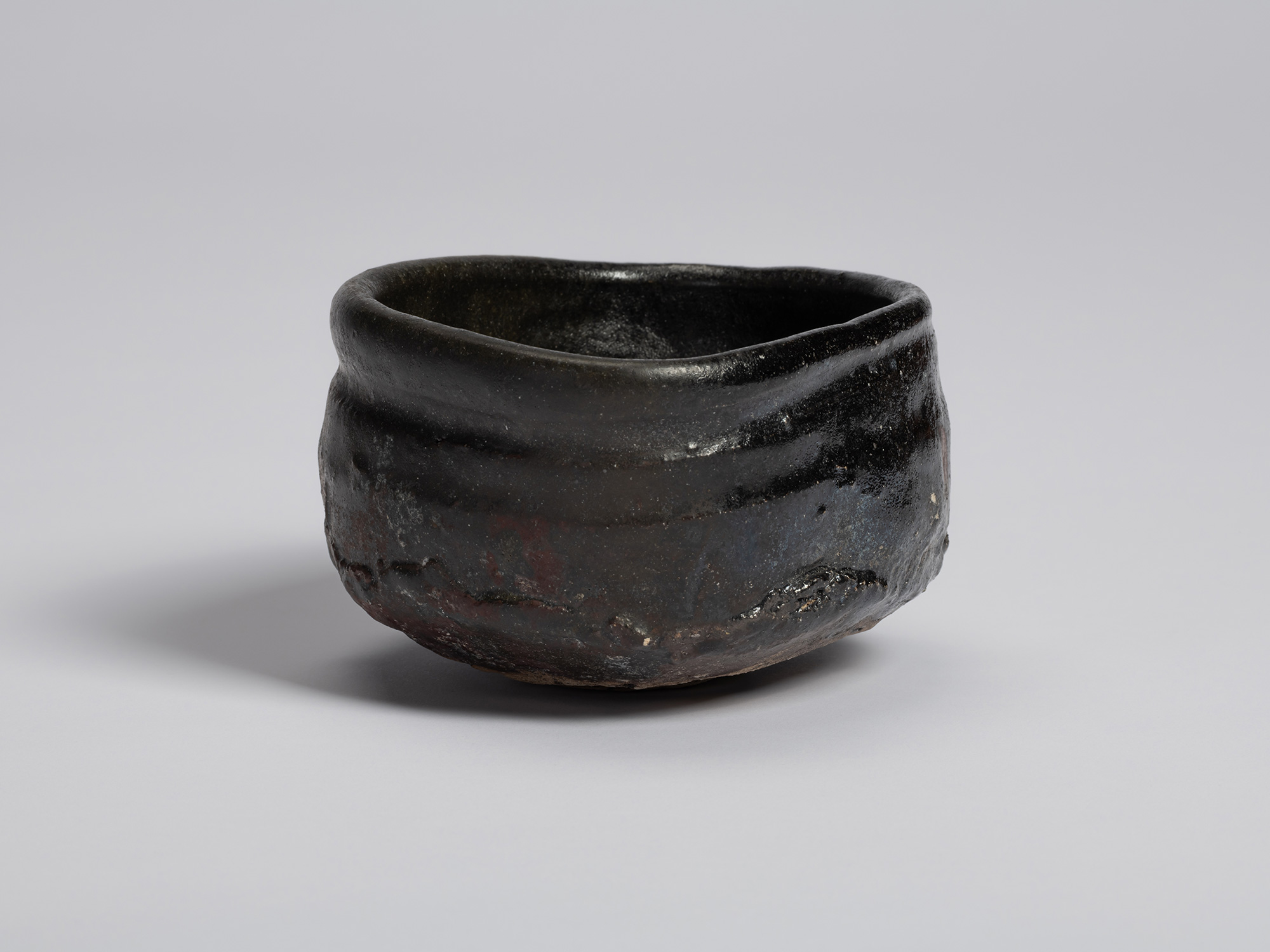View of a black tea bowl by Shiro Tsujimura. The bowl's irregular rim rises and dips around the...