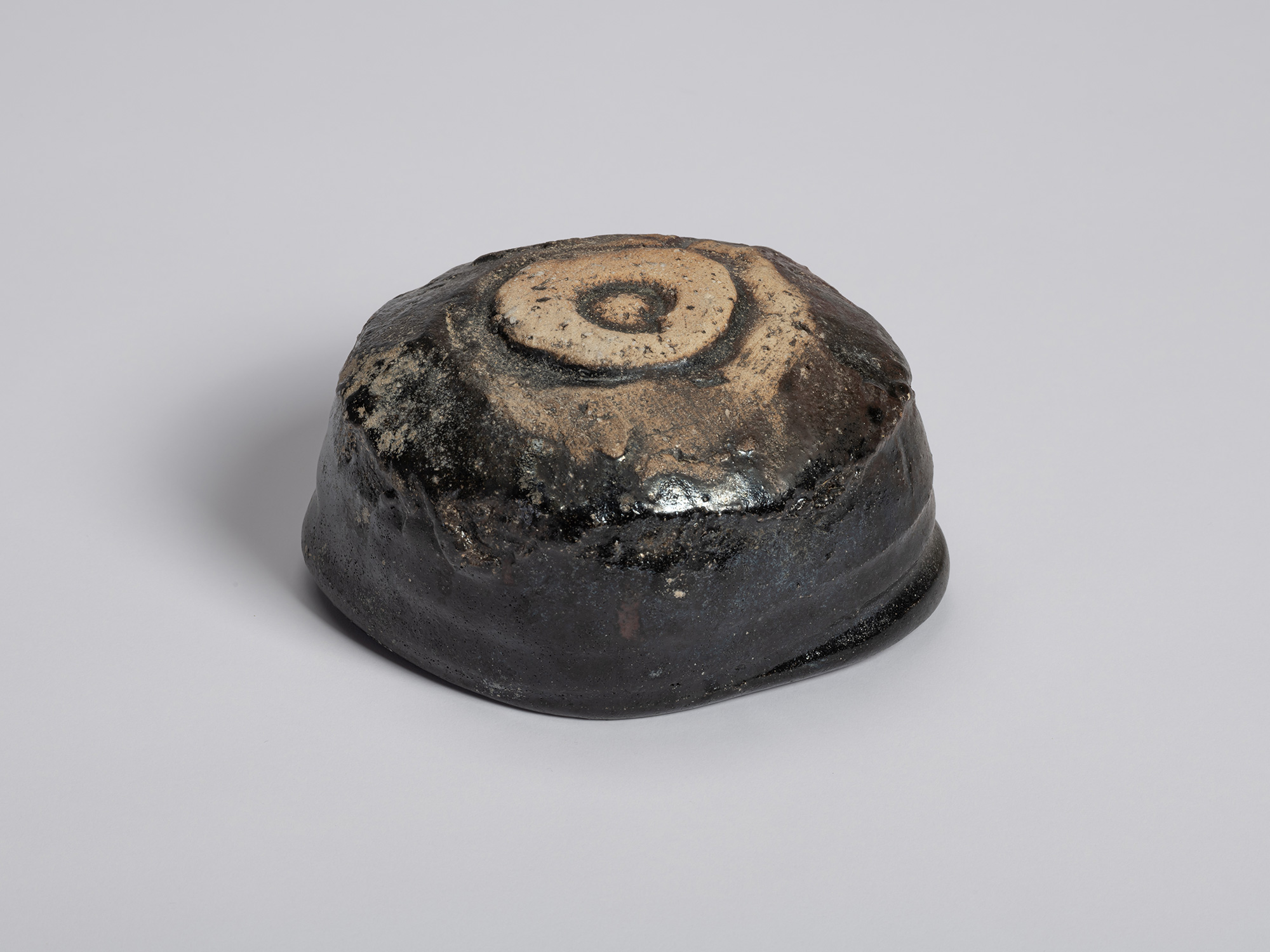 Three-quarter view of the underside of a black tea bowl by Shiro Tsujimura. The bowl rests on an...