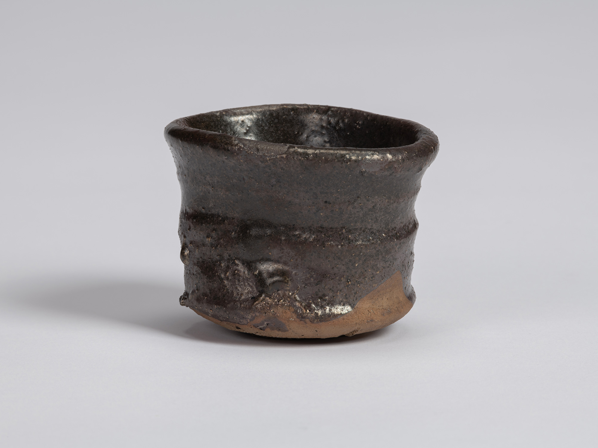 Kuro Guinomi by Shiro Tsujimura, with an irregular cylindrical form, dark black-brown glazed surface,...