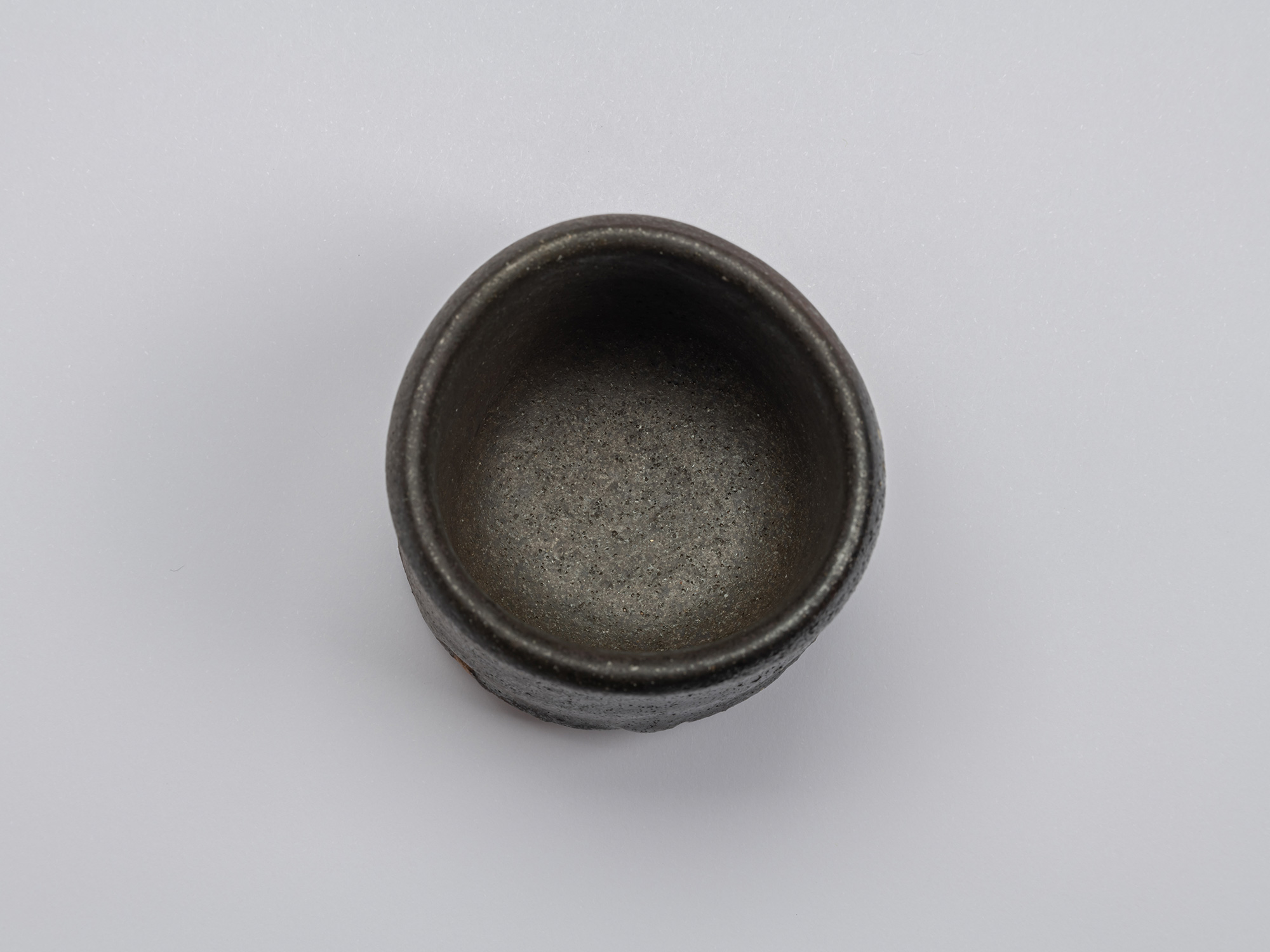 Overhead view of Shiro Tsujimura’s kuro guinomi showing a rounded irregular rim and dark, finely...