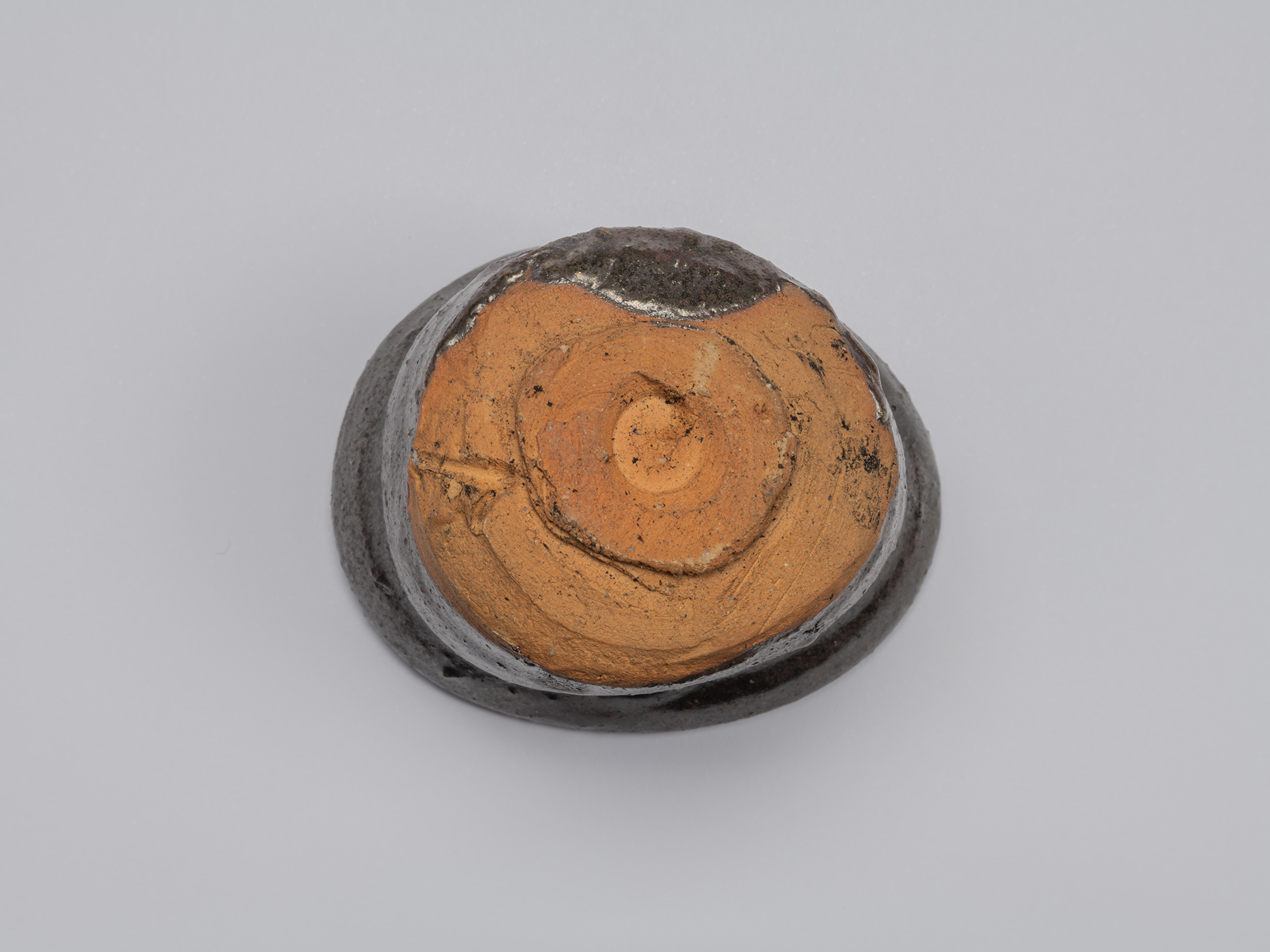 Underside of Shiro Tsujimura’s kuro guinomi showing an unglazed reddish-brown clay base, roughly...