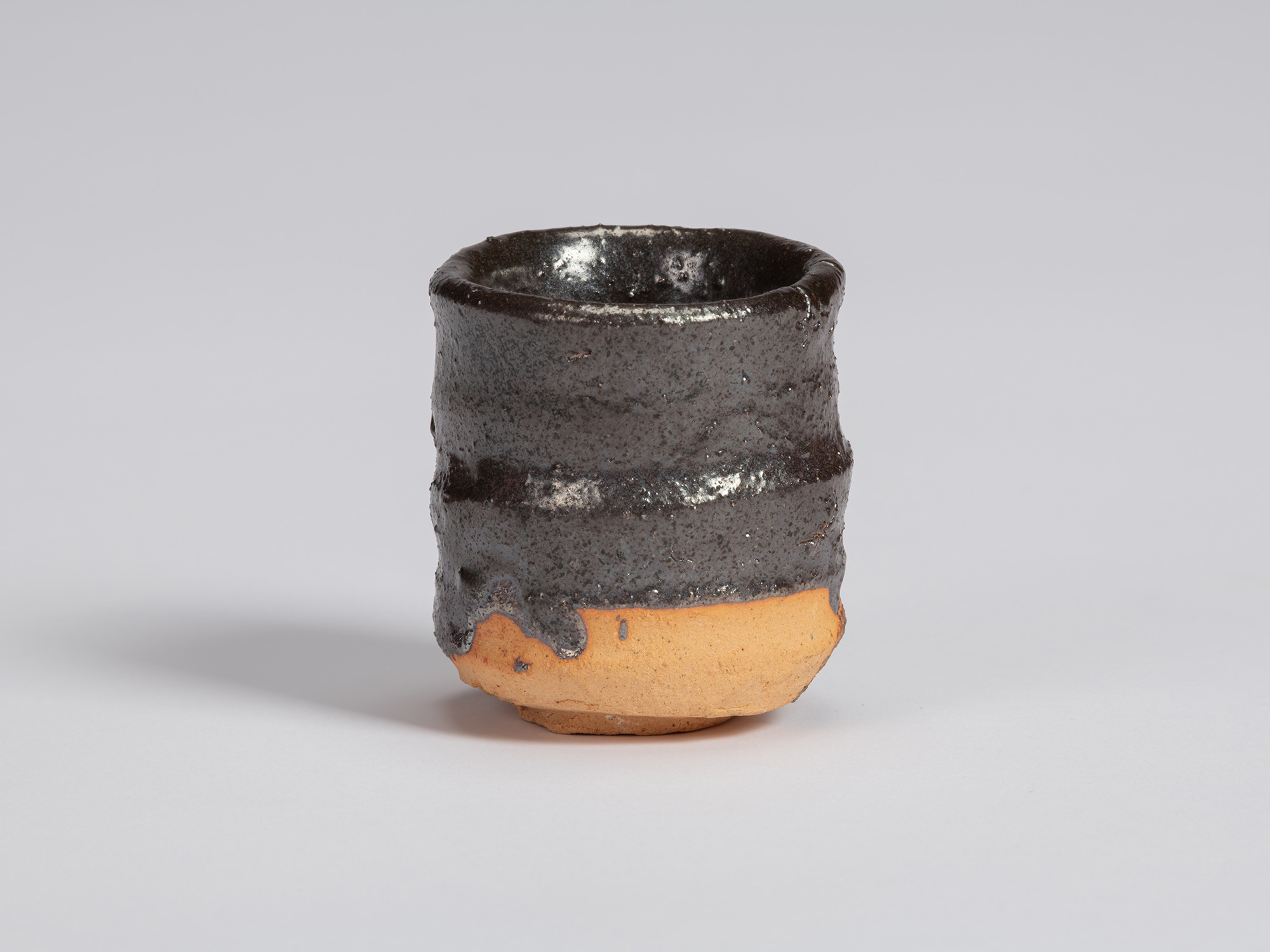Kuro Guinomi by Shiro Tsujimura, with an irregular cylindrical form, glossy black glaze, and areas...