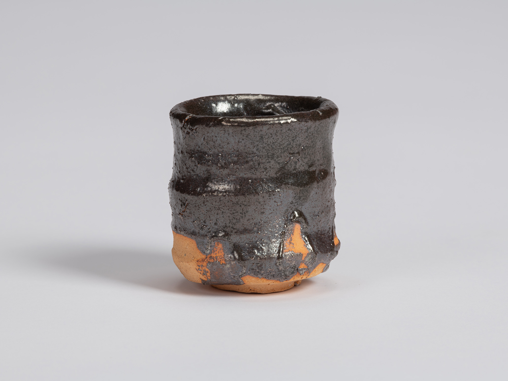 Kuro Guinomi by Shiro Tsujimura, with an irregular cylindrical form, glossy black glaze, and areas...