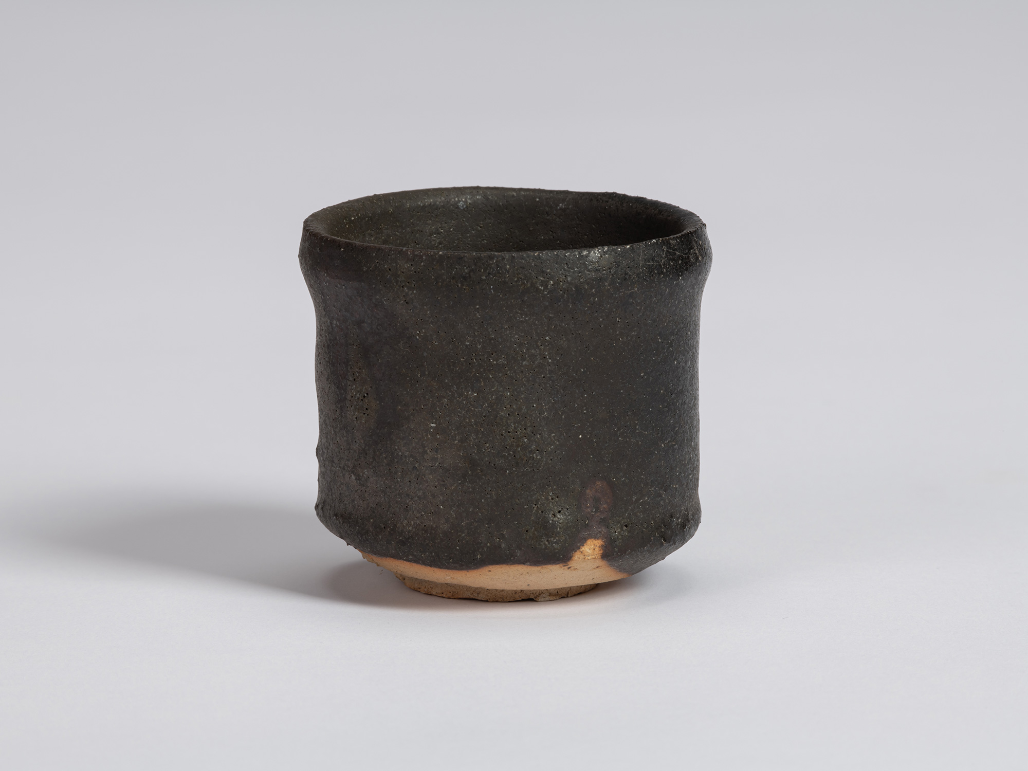 Kuro Guinomi by Shiro Tsujimura, with a tall cylindrical form, matte black-brown surface, subtly...