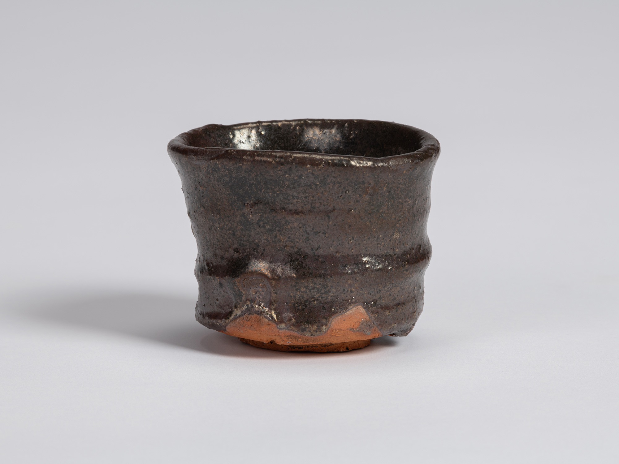 Kuro Guinomi by Shiro Tsujimura, with a cylindrical form, irregular rounded rim, and dark brown-black...