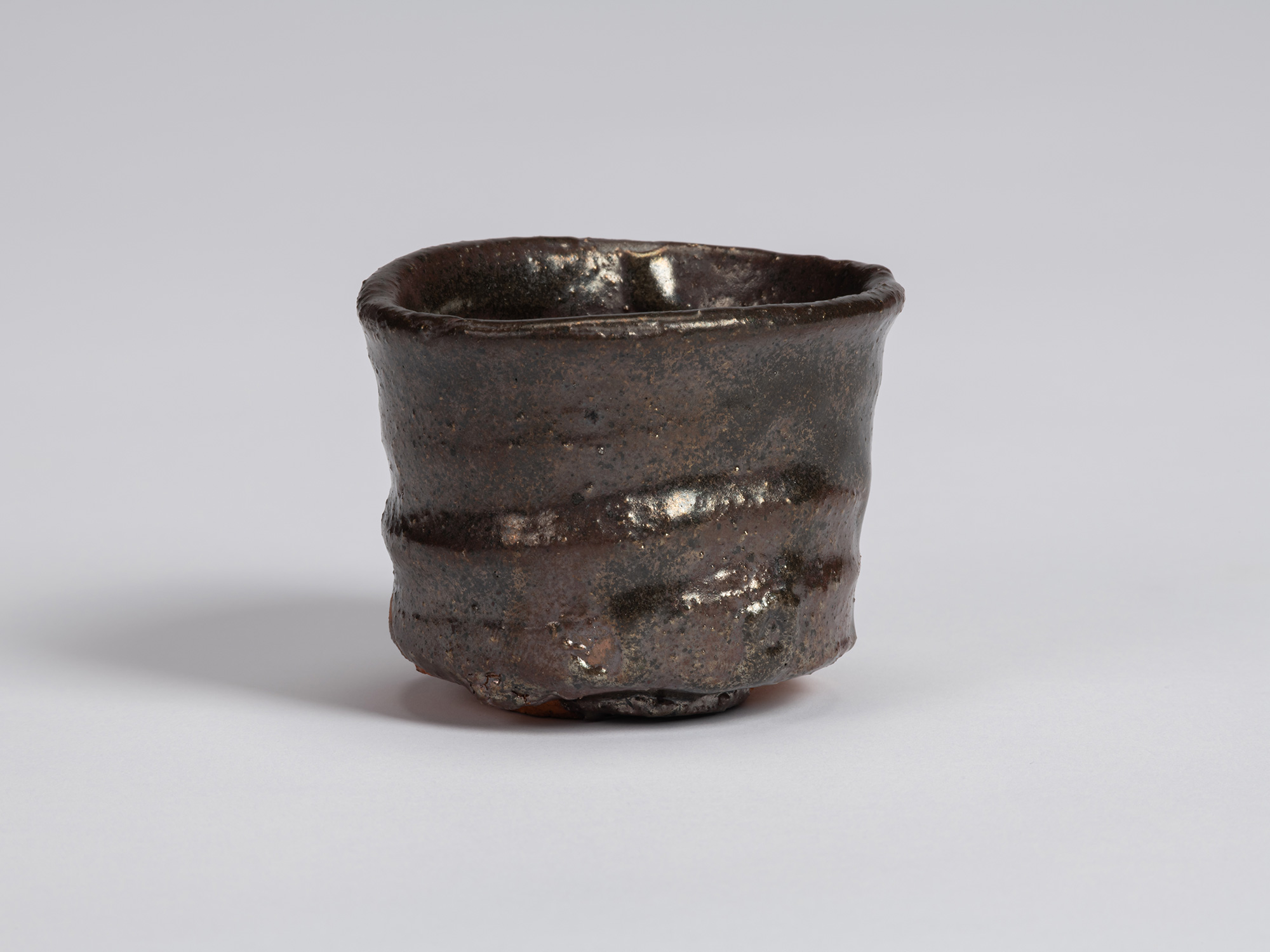 Kuro Guinomi by Shiro Tsujimura, with a cylindrical form, irregular rounded rim, and dark brown-black...