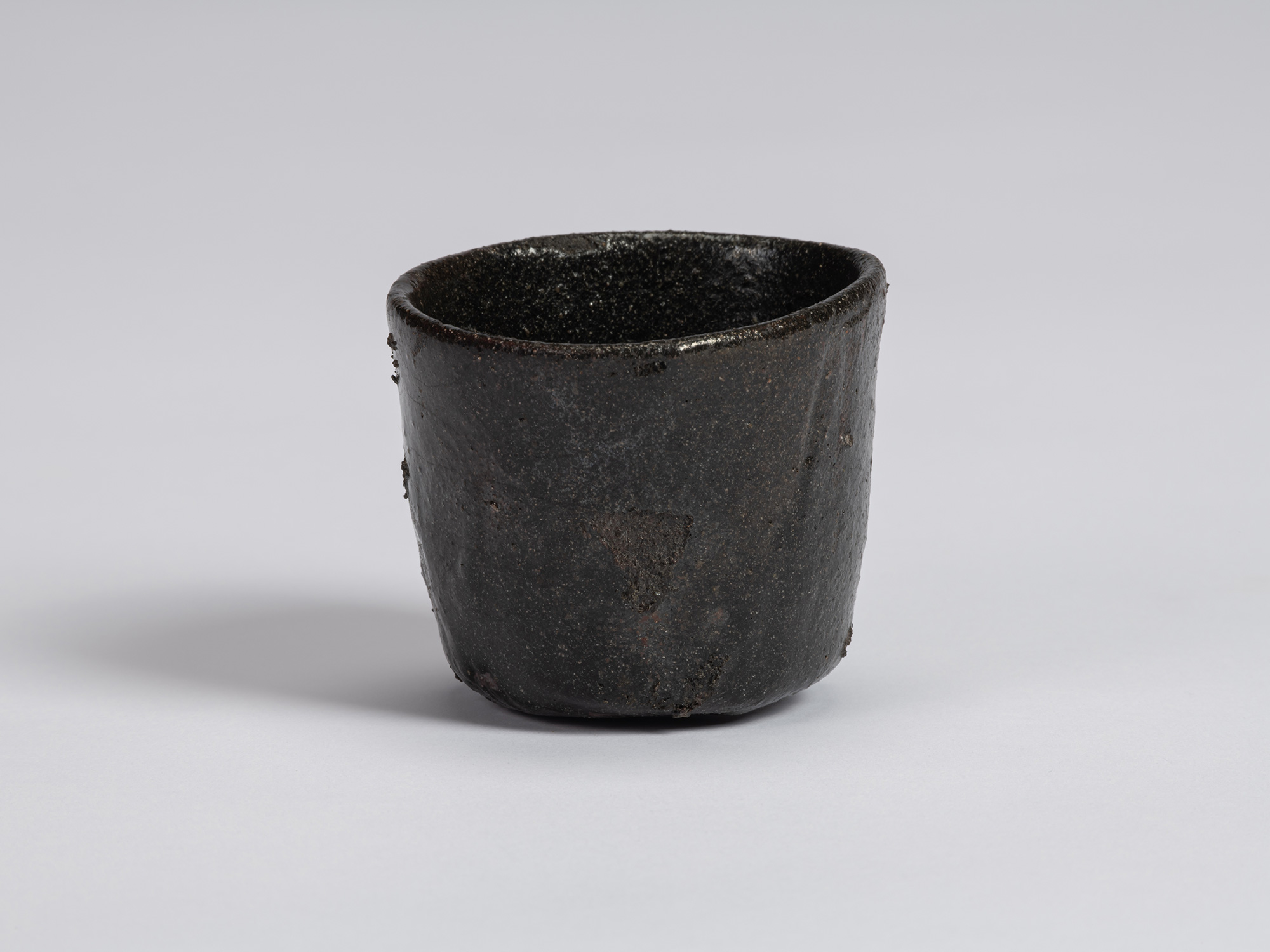 Kuro Guinomi by Shiro Tsujimura, front view, with a slightly tapered form, irregular rim, and textured...