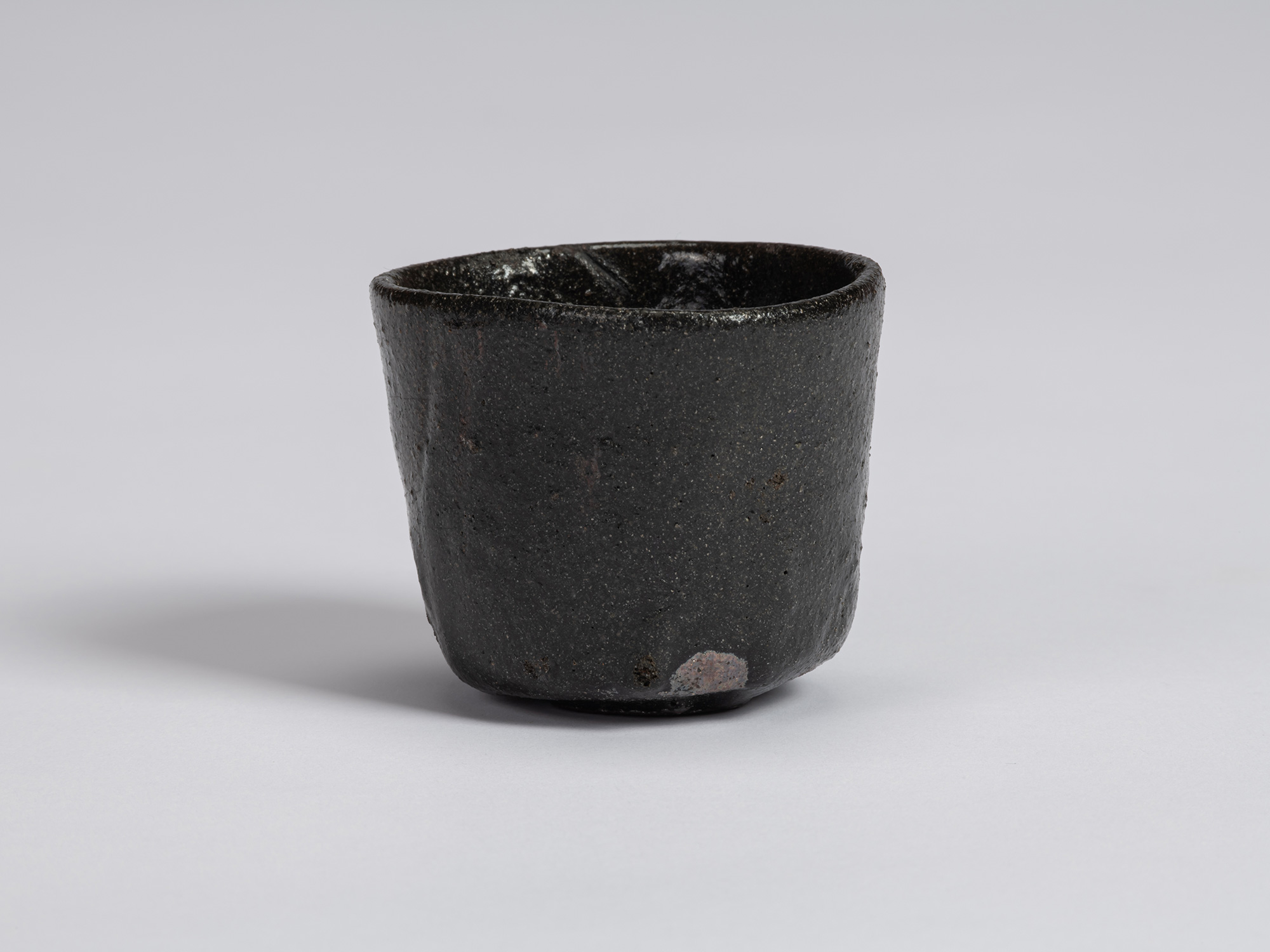 Kuro Guinomi by Shiro Tsujimura, front view, with a slightly tapered form, irregular rim, and textured...