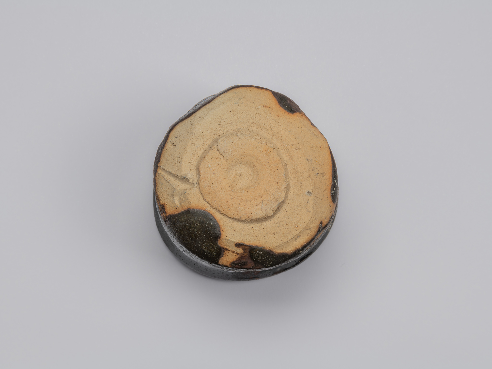 Underside of Shiro Tsujimura’s Kuro Guinomi, showing a broad expanse of unglazed tan clay, roughly...